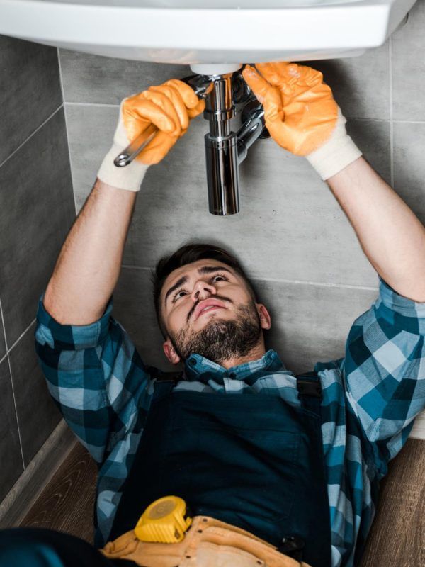 Plumber in orange gloves, lying on floor, fixing sink pipes with a wrench.