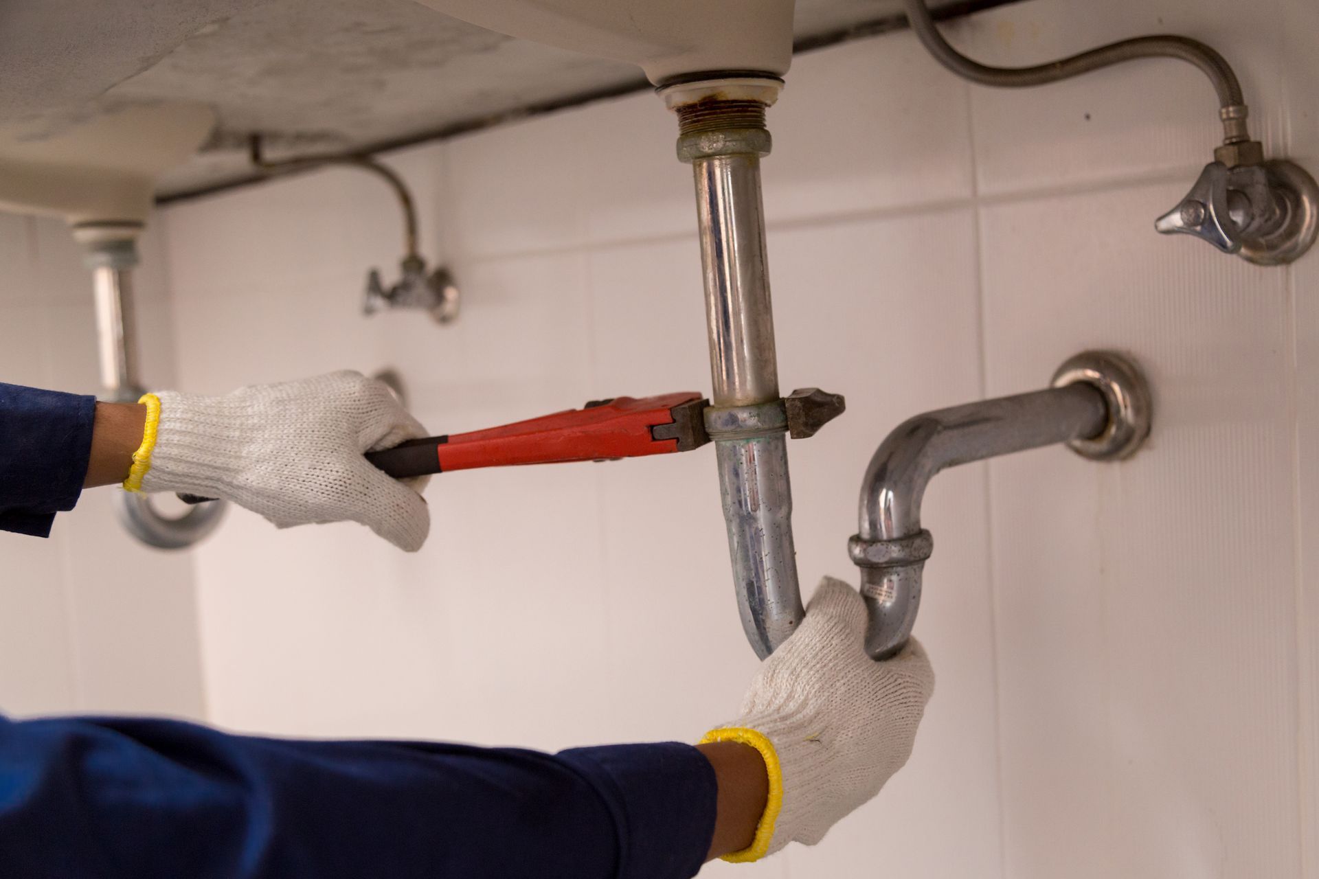 Plumber using a wrench to repair a curved drainpipe under a white sink in a tiled room.