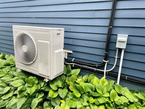 Air conditioning unit outside a blue house, surrounded by green bushes. Electrical conduit visible.