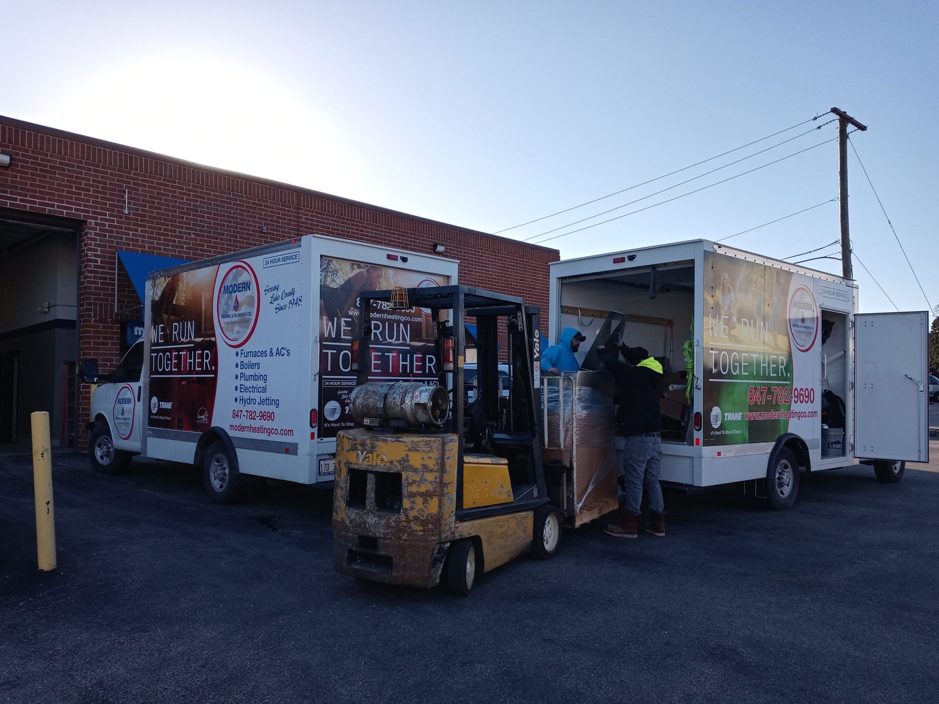 A forklift operator loads cargo into a truck next to another truck; both vehicles have company logos.