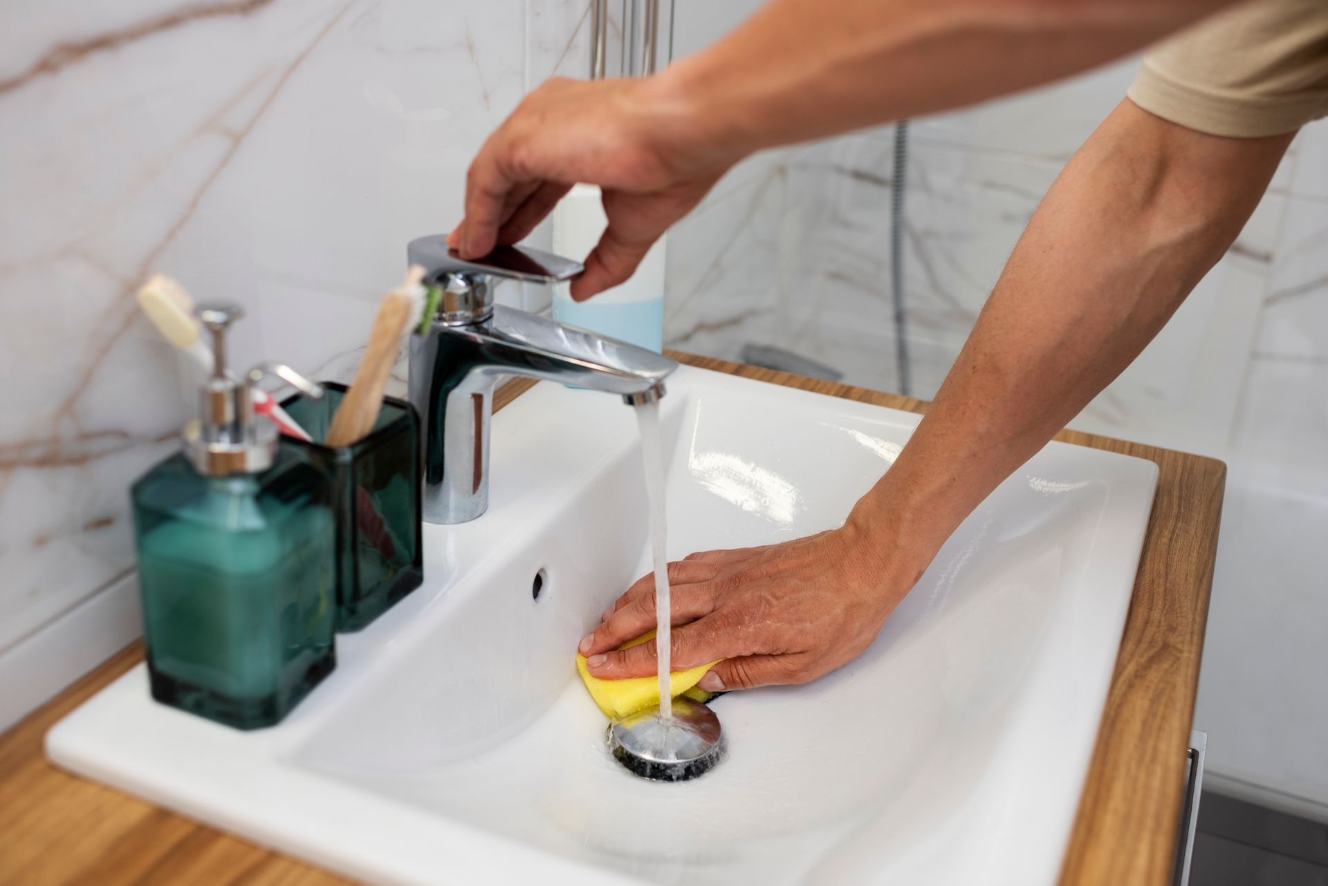 Person cleans a white bathroom sink with a yellow sponge under running water.