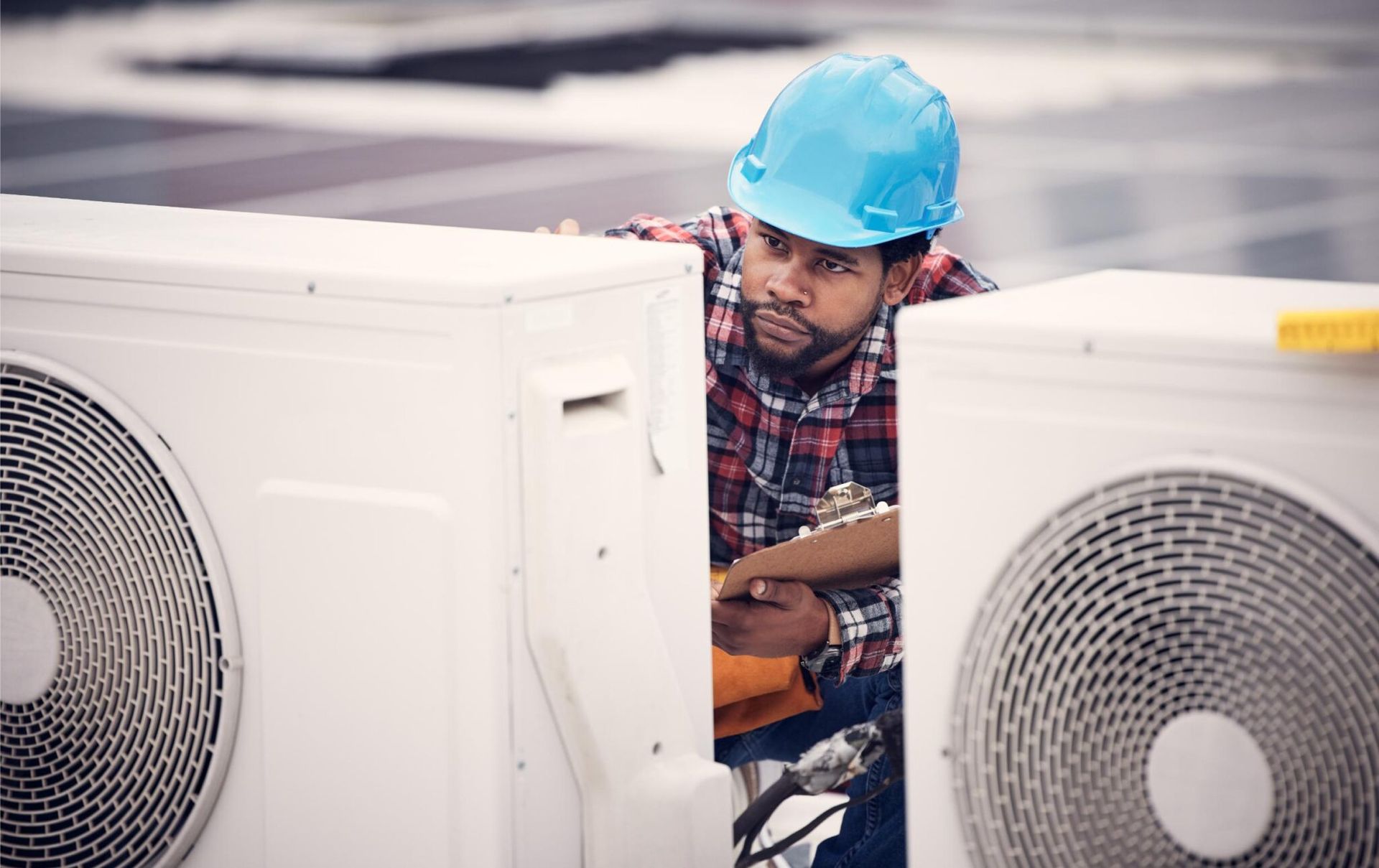 HVAC technician inspecting air conditioning units on a rooftop, holding a clipboard and wearing a blue hard hat.
