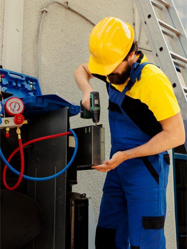 HVAC technician in yellow hard hat and blue overalls using a drill outdoors, near equipment and a ladder.
