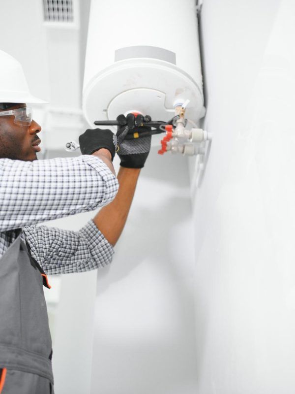 Plumber in a hard hat and gloves working on a water heater with a wrench. White wall background.