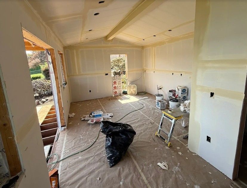 Room under construction with drywall, tape, paint buckets, and debris on the floor.