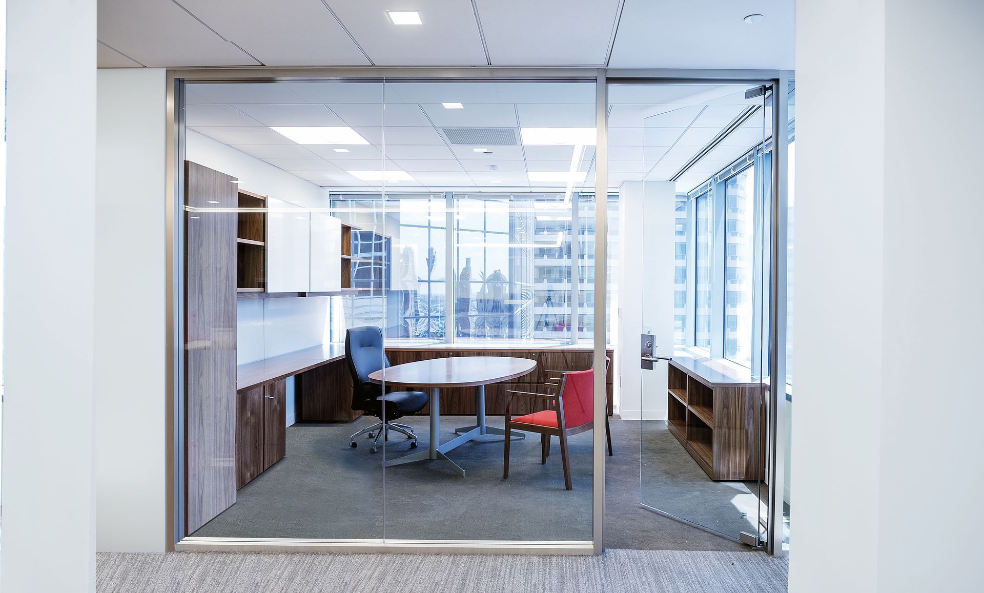 Glass-walled office with a round table, chairs, built-in cabinets, and a city view.