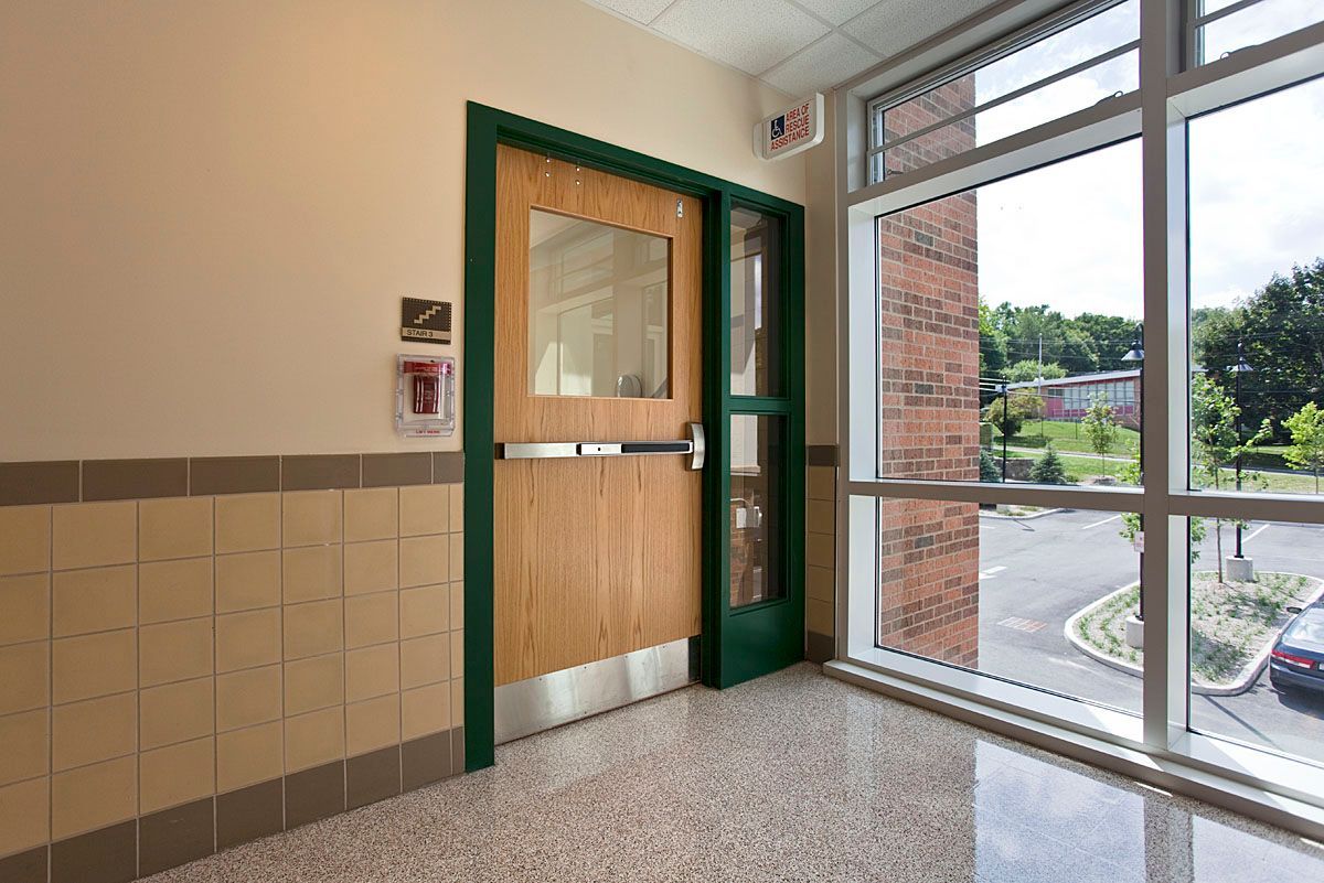 Doorway with window, brick wall, and fire alarm in school hallway.