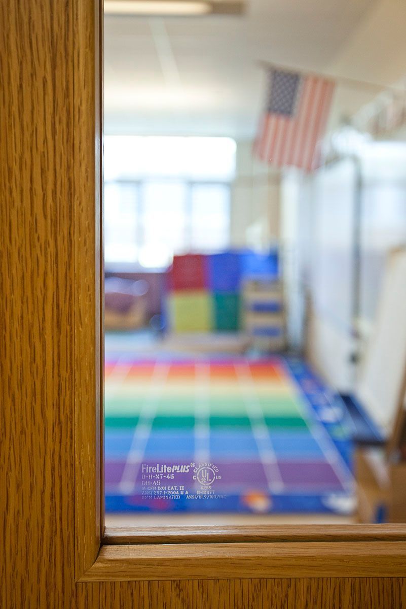 Wooden door with window overlooking a classroom with a rainbow-colored floor mat, an American flag, and colorful blocks.