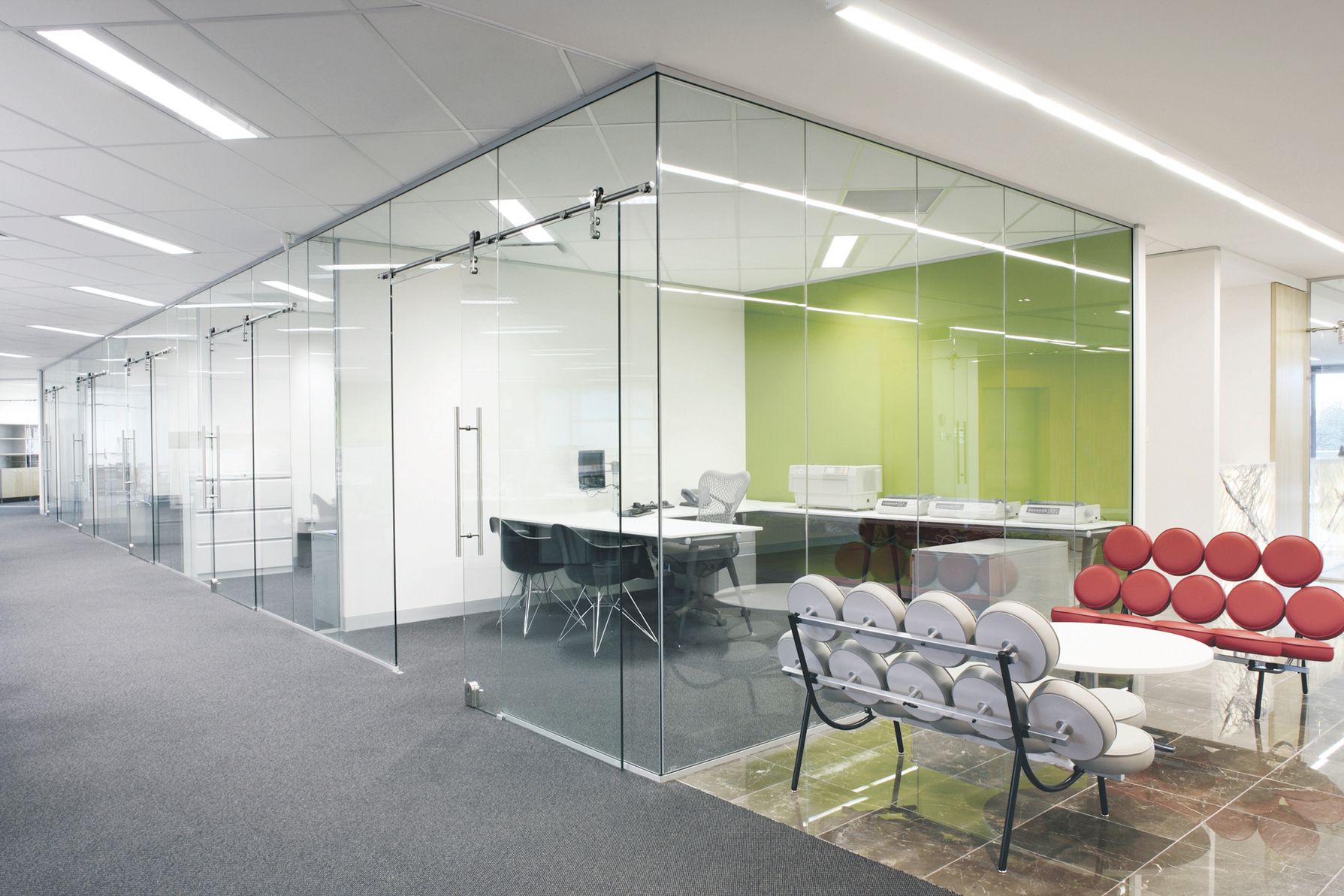 Modern office space with glass walls, desks, and chairs. Green accent wall and red sofa.