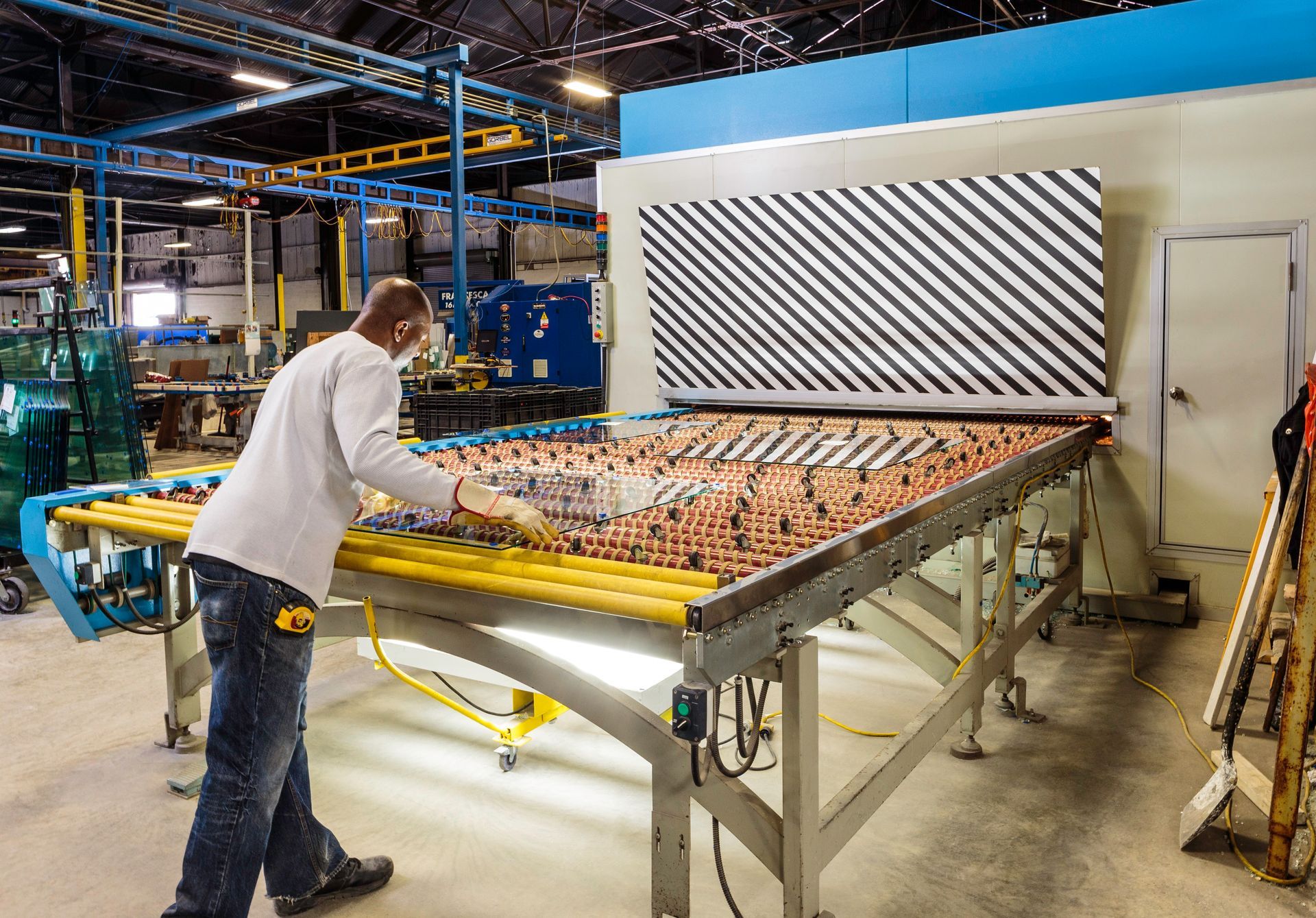 Worker in factory inspecting glass on conveyor belt in front of a machine.