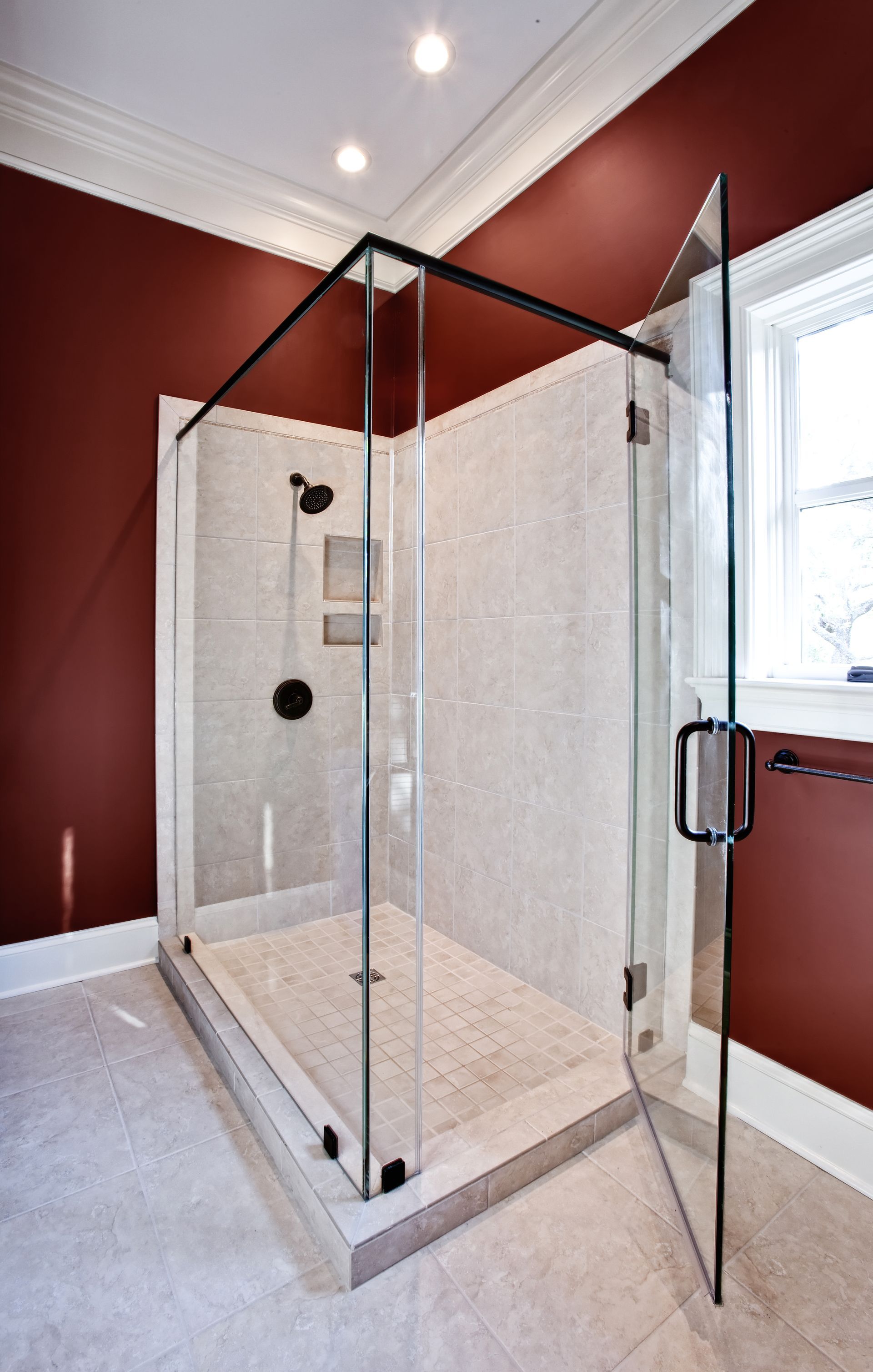 Glass shower with open door in a bathroom with red walls, white trim, and a window.