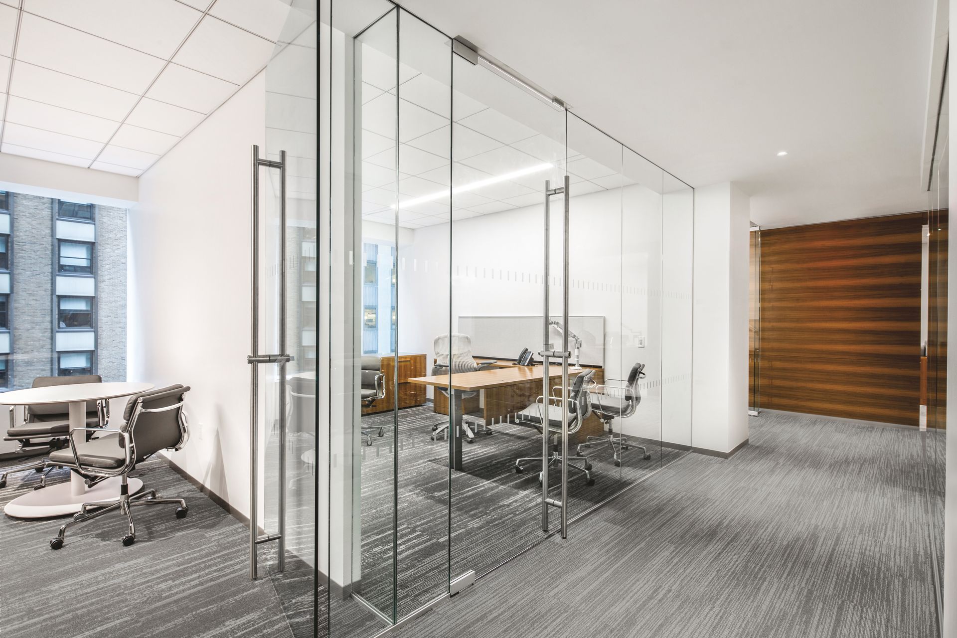 Glass-walled office space with a desk, chairs, and wooden door, carpeted floor, and natural light.