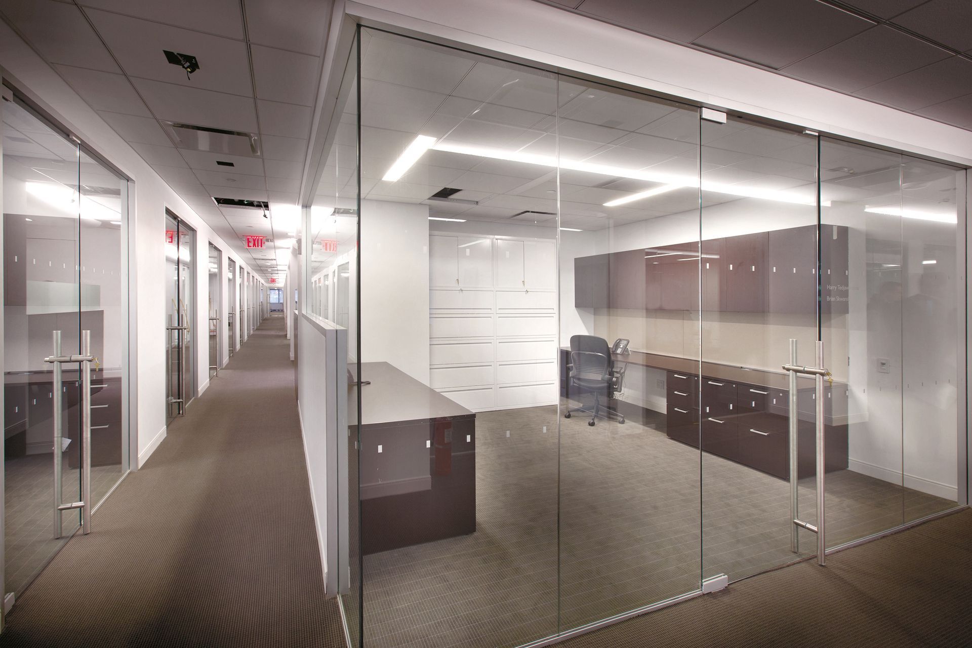 Office hallway with glass-walled rooms, brown carpet, and overhead lighting.