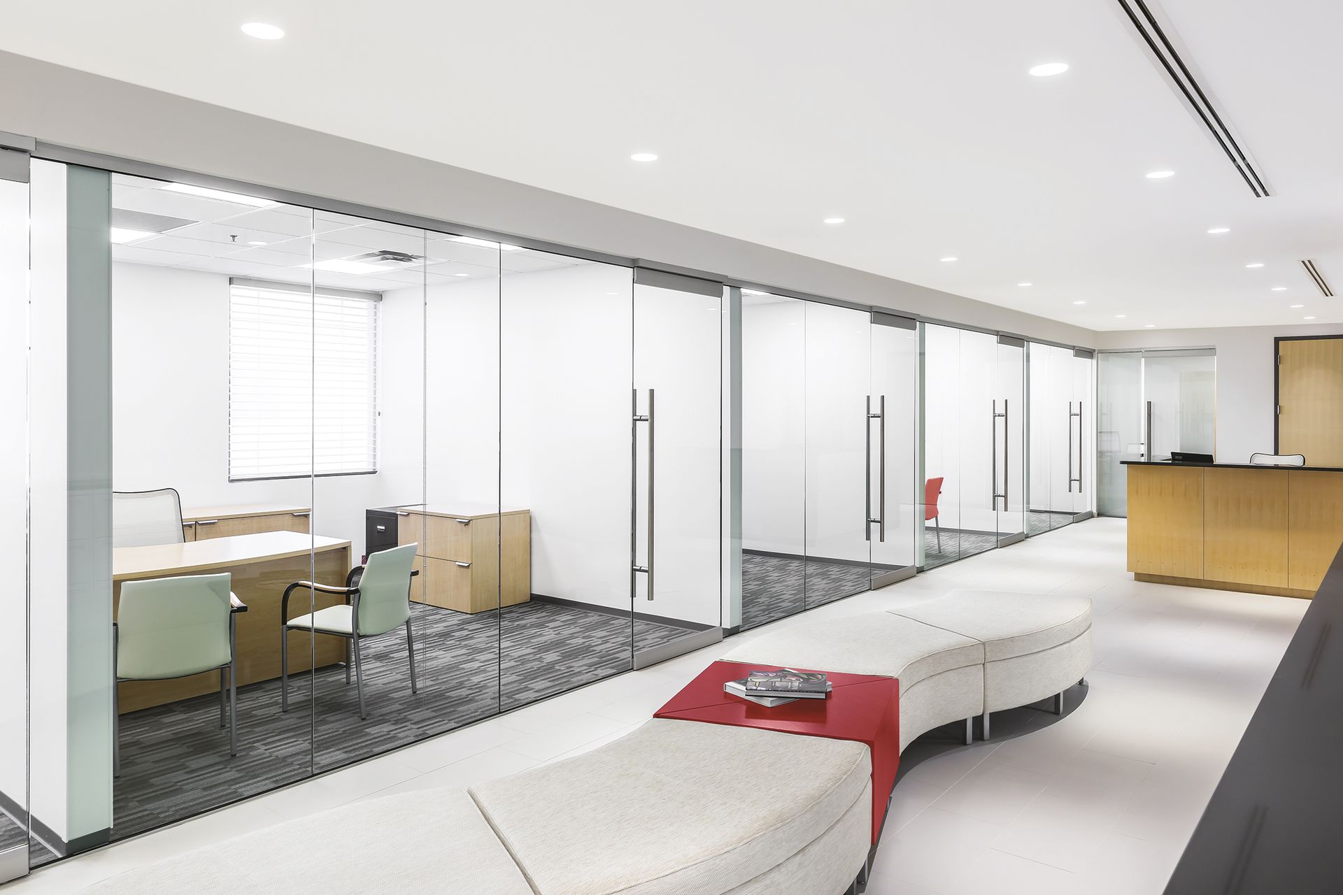 Modern office hallway with glass-walled rooms, a curved bench, and a red side table.