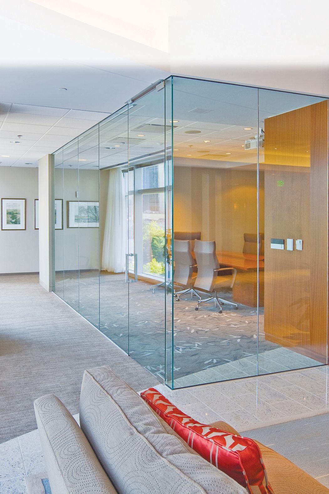 Glass-walled office with a view of a desk, chairs, and a hallway with a sofa and artwork.