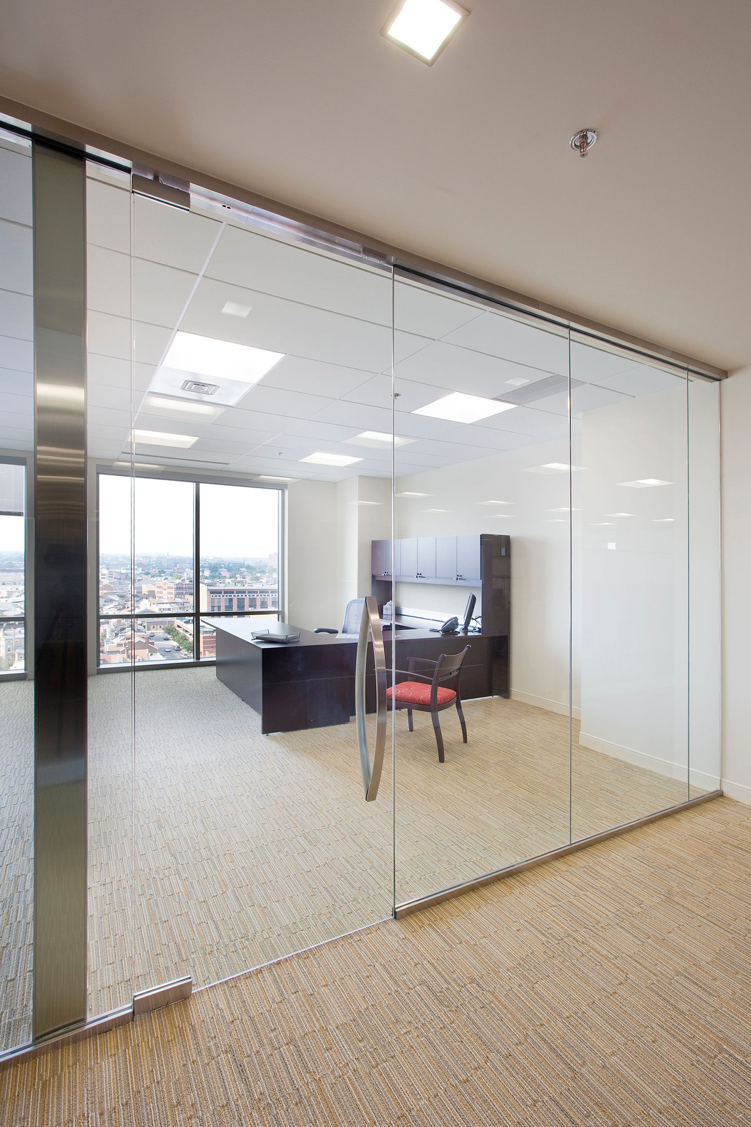 Glass-walled office with desk, chair, and city view. Beige carpet.