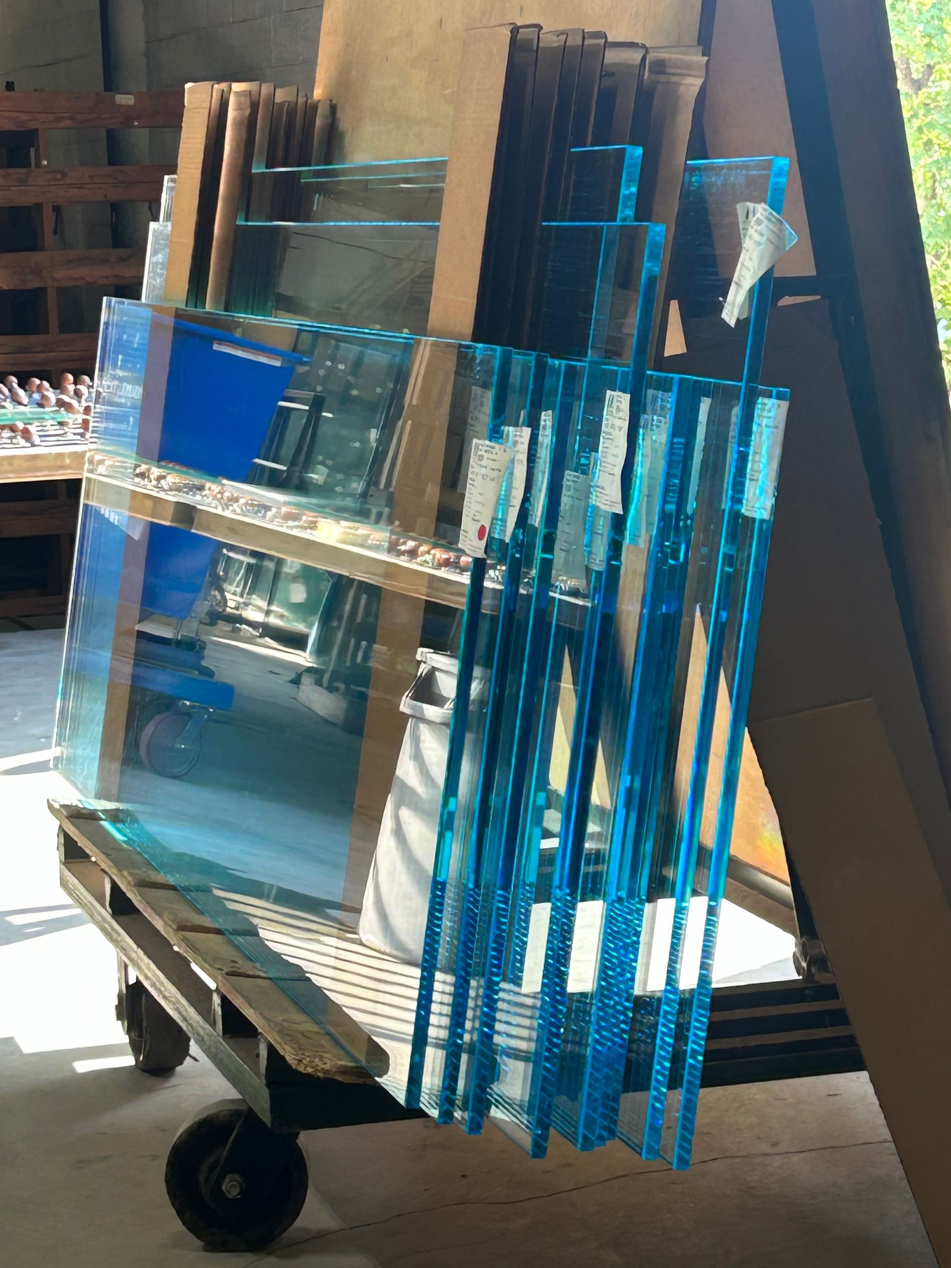 A cart holds stacks of clear glass sheets, some with blue protective covering, in a warehouse.