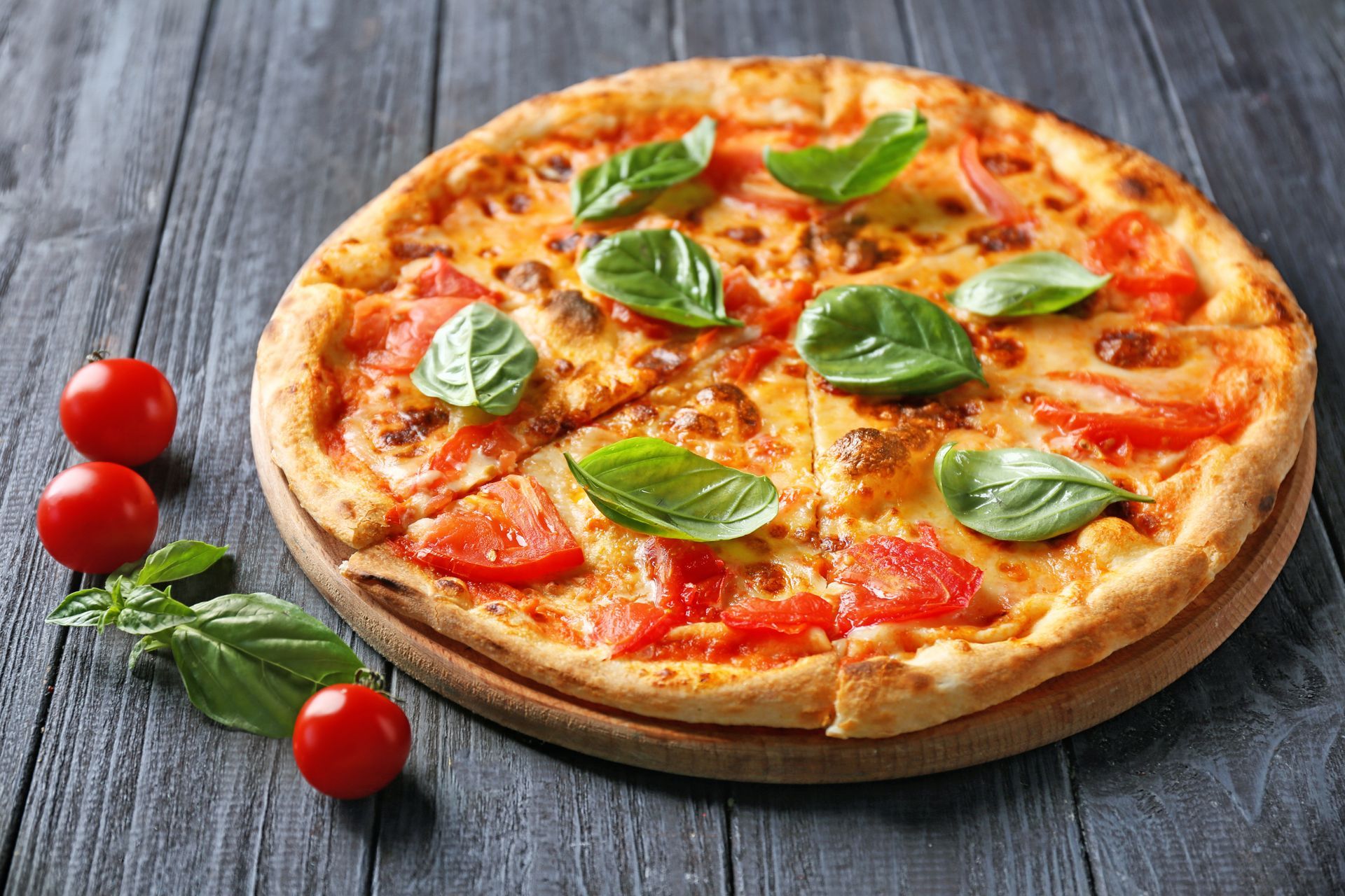 Pizza topped with tomatoes and basil on a wooden board with cherry tomatoes next to it.