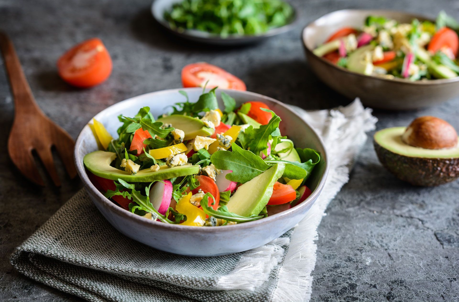 Salad bowl with avocado, tomatoes, greens, and yellow pepper, on a gray surface with additional ingredients.
