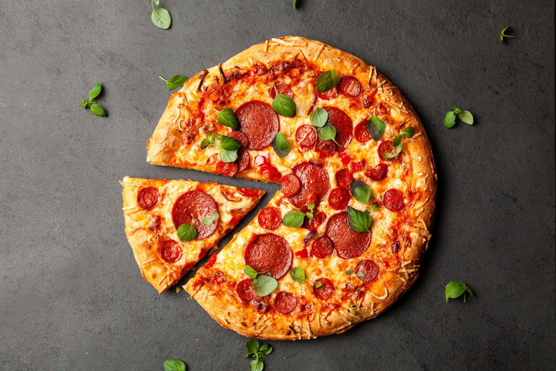 Pepperoni pizza with a slice cut out, garnished with green herbs, on a dark surface.