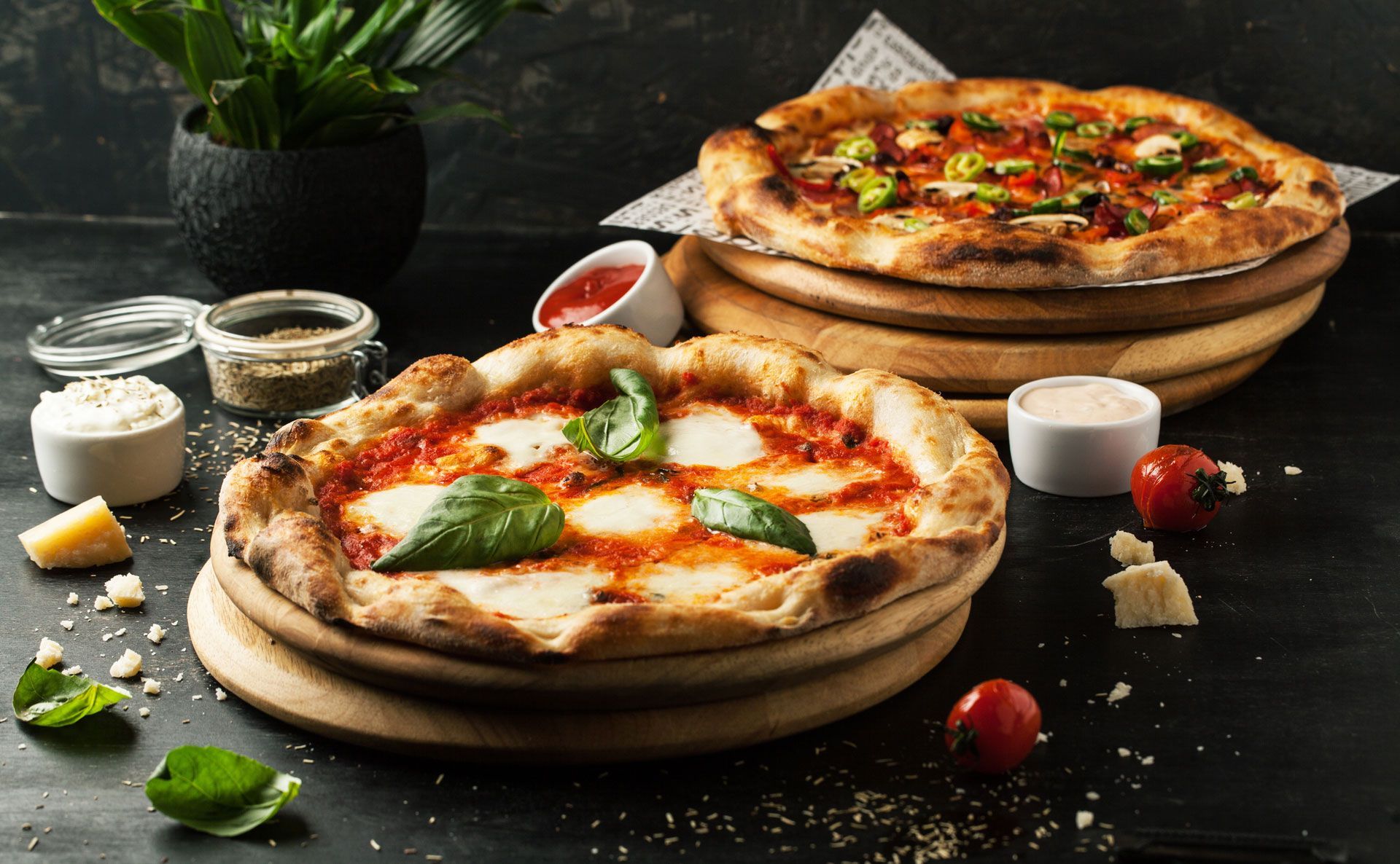 Two pizzas with various toppings on wooden platters, surrounded by ingredients and sauces.