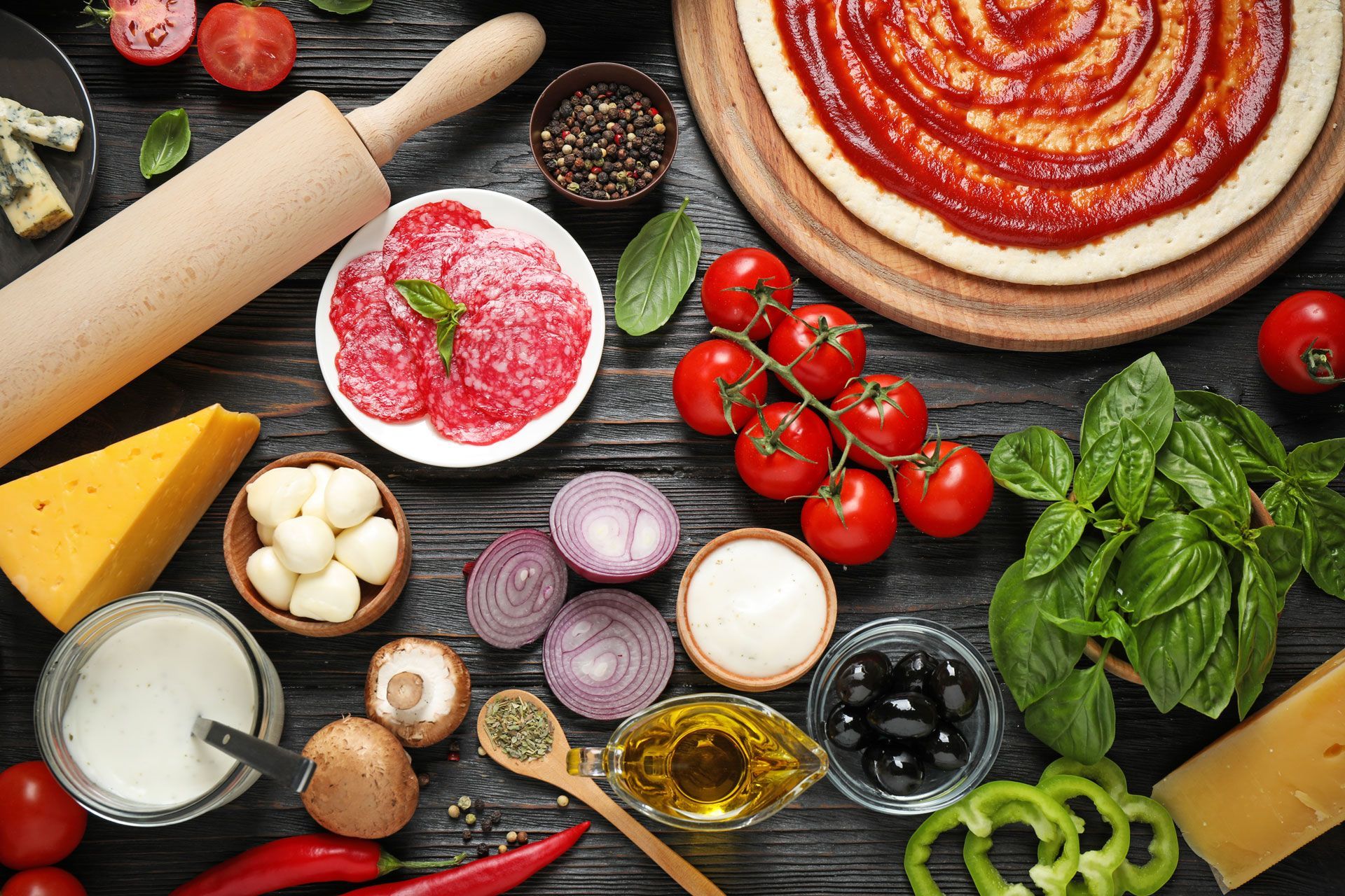 Pizza ingredients laid out on a dark wooden surface: dough, sauce, cheese, tomatoes, salami, herbs, and vegetables.