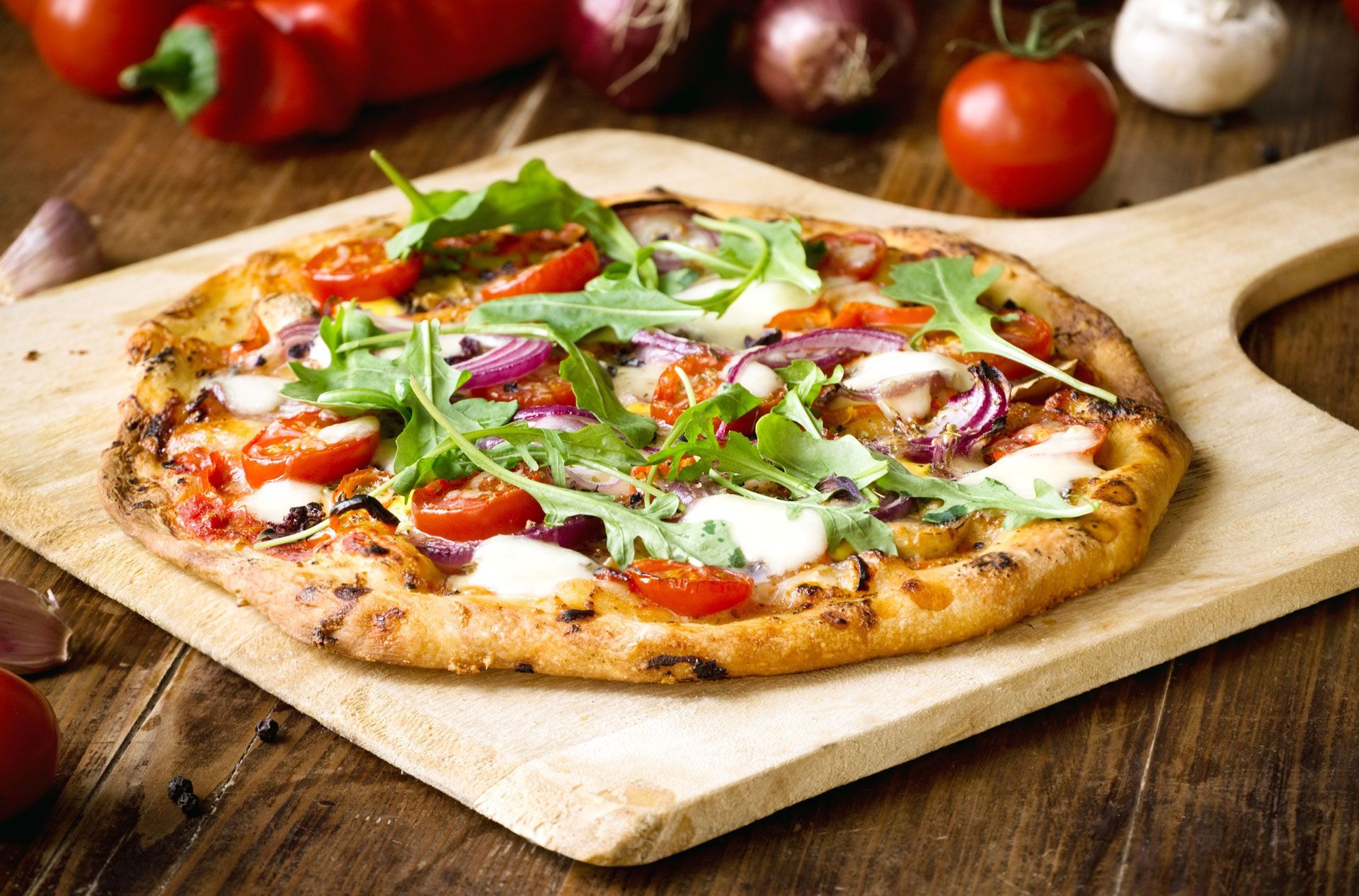 Pizza on a wooden board, topped with tomatoes, red onion, and arugula.