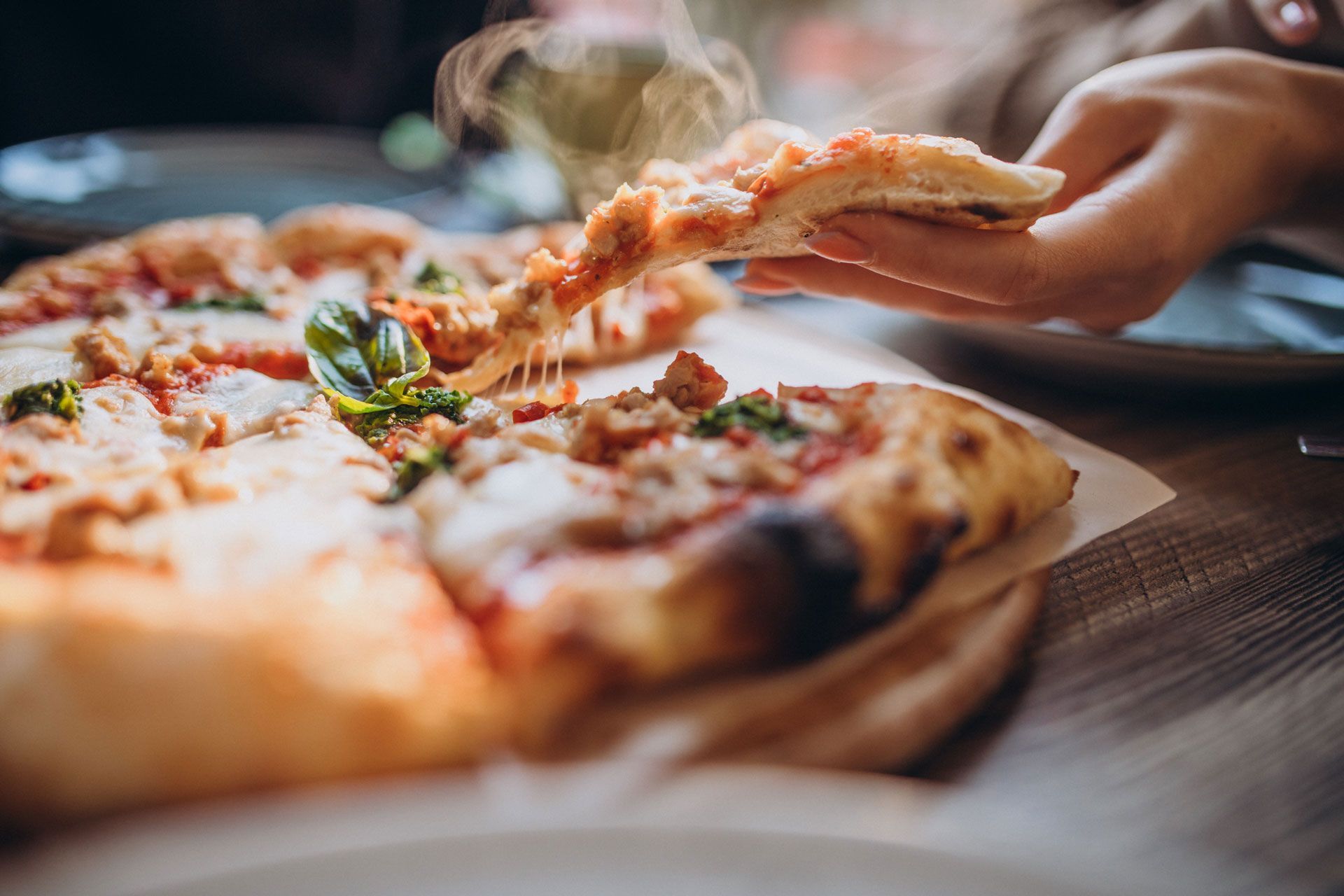 Pizza slice being lifted from a whole pizza on a wooden table, steam rising, hand visible. Pizza slice being lifted from a whole pizza on a wooden table, steam rising, hand visible.