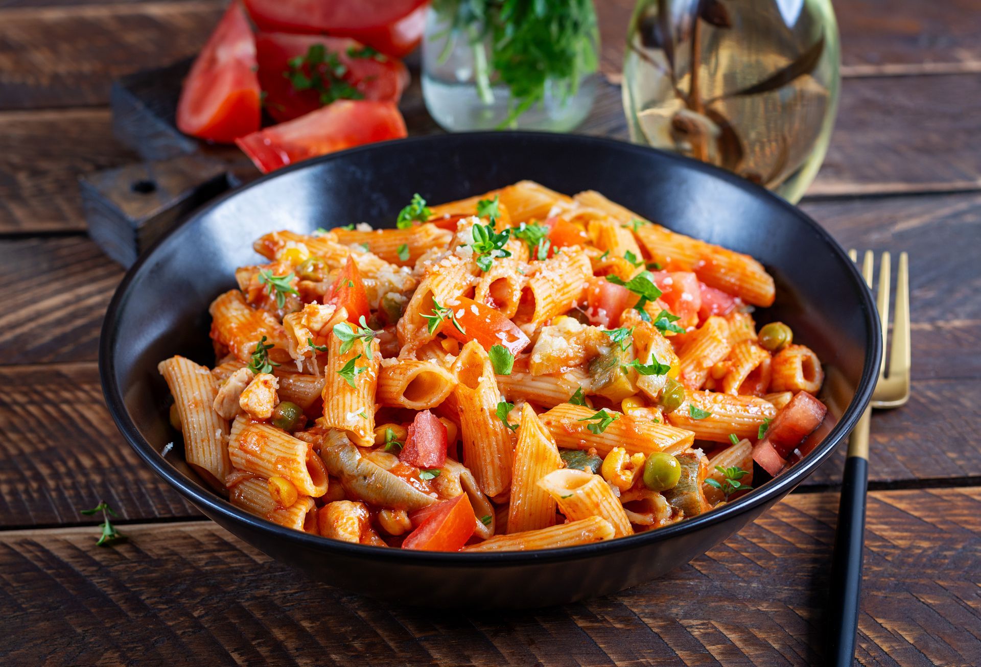 Penne pasta dish with tomatoes, peas, and parsley in a black bowl on a wooden table.
