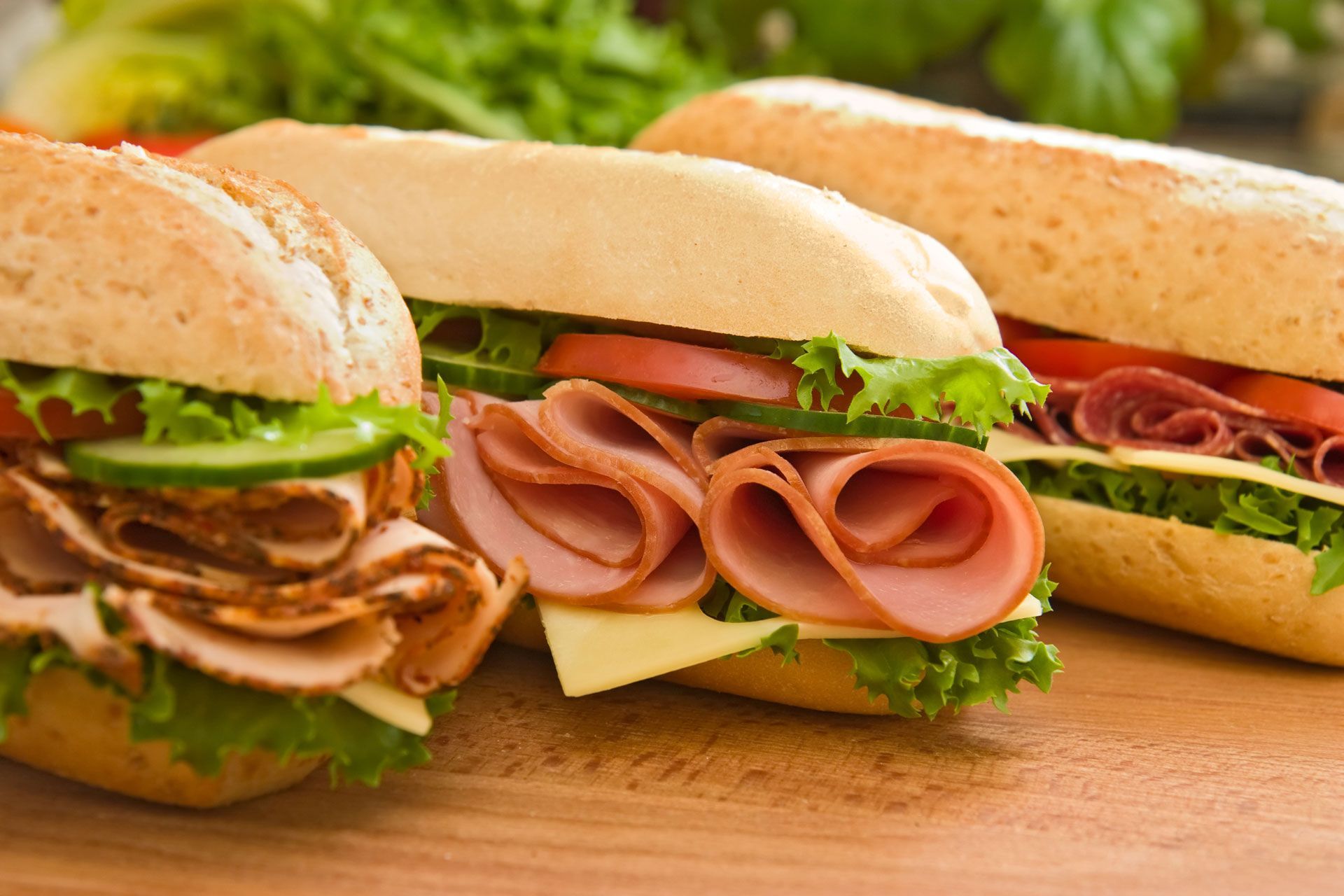 Three sandwiches with meat, cheese, lettuce, and tomatoes on a wooden surface.