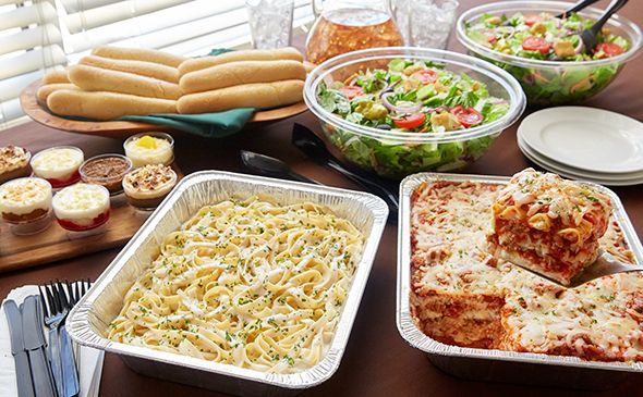 Table spread with catered Italian food including pasta, lasagna, breadsticks, salad, and desserts.