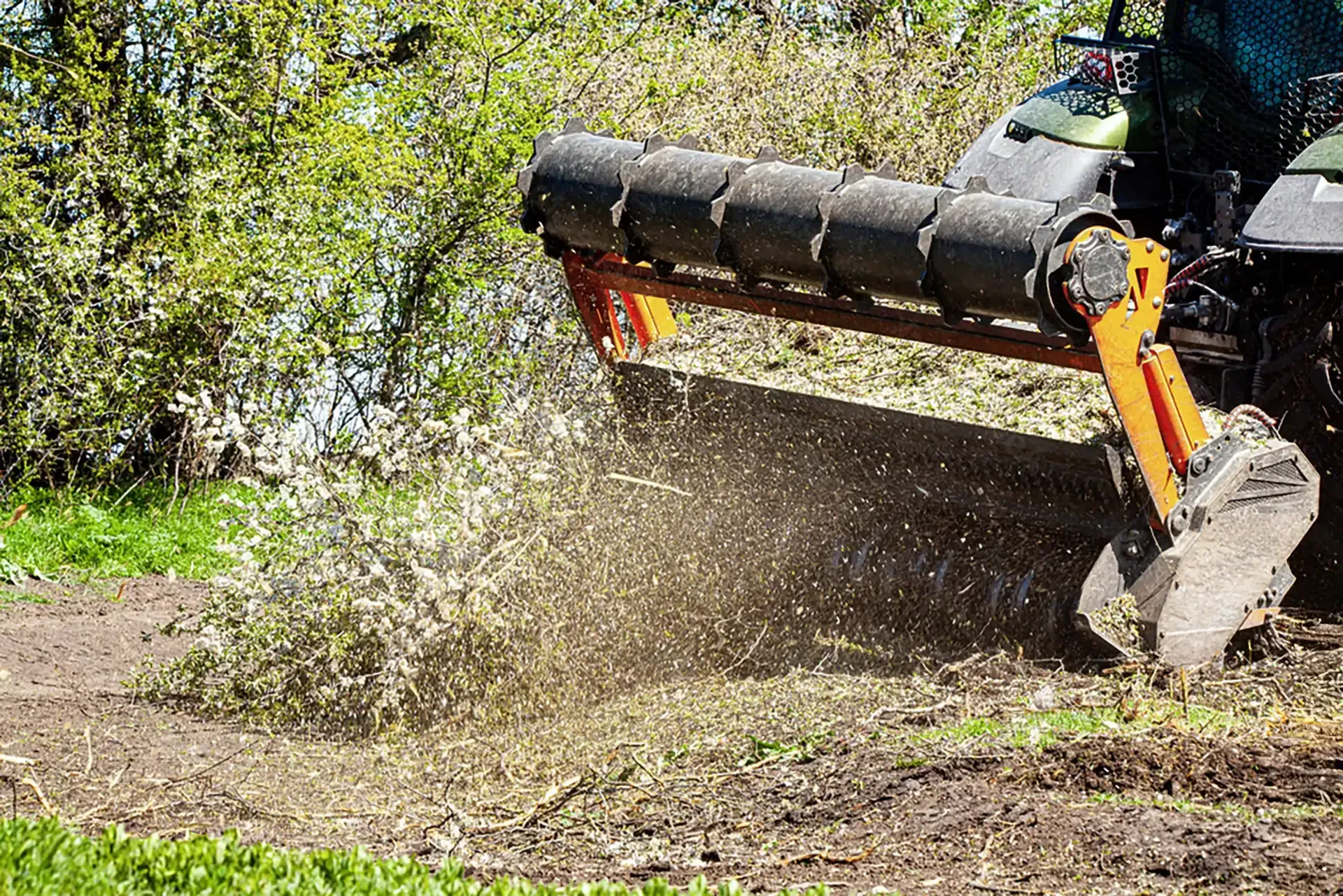 A tractor mulching brush and small trees, kicking up wood chips. The scene is outdoors with green foliage and brown earth.