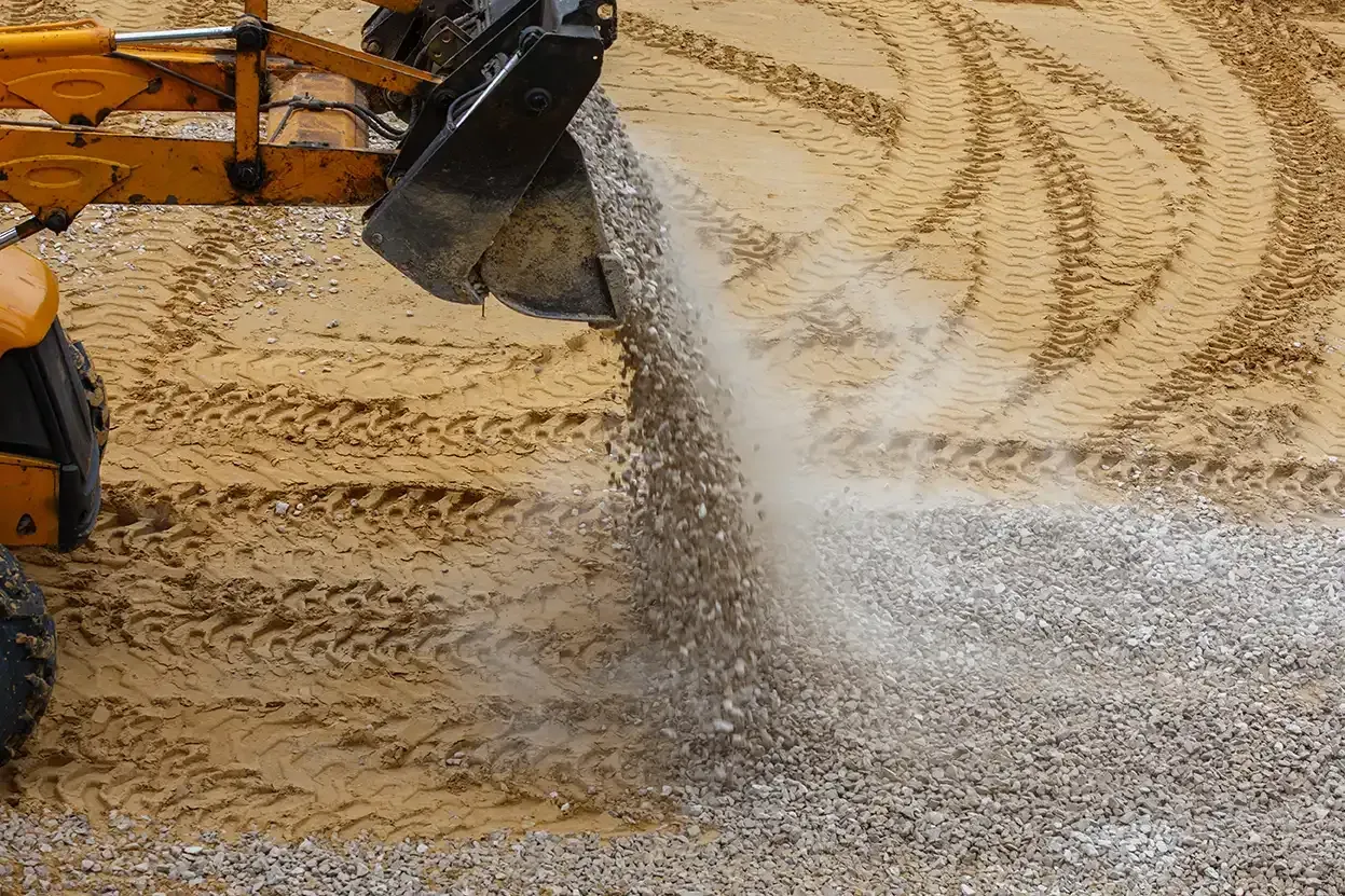 A front-end loader is spreading gravel on a dirt surface, creating a dust cloud.