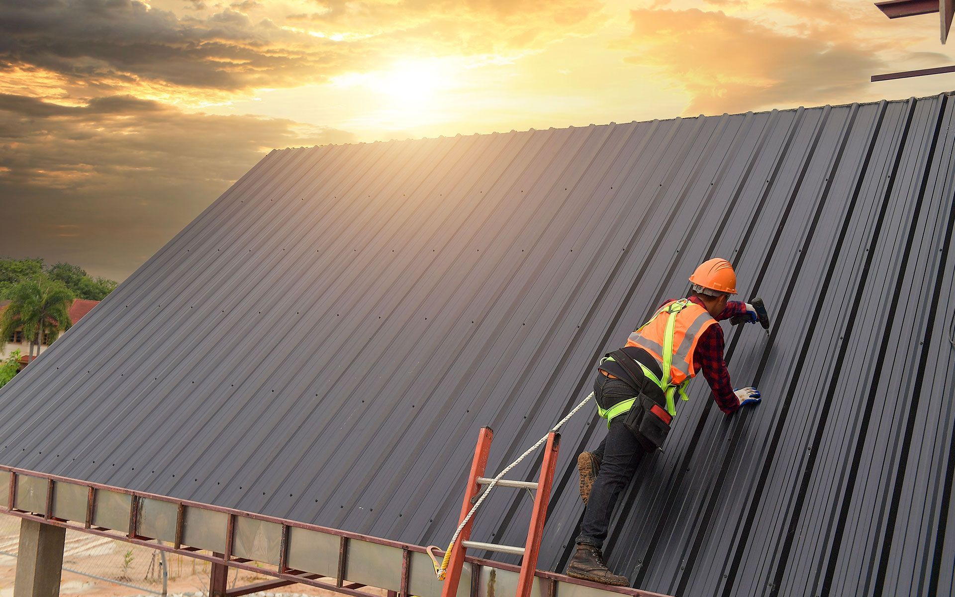 Roofer in safety gear working on a metal roof under a sunset sky.