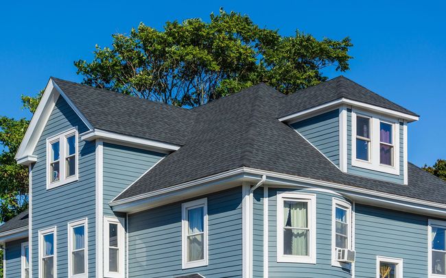 Blue house with dark roof and white trim against a blue sky, a leafy tree visible.