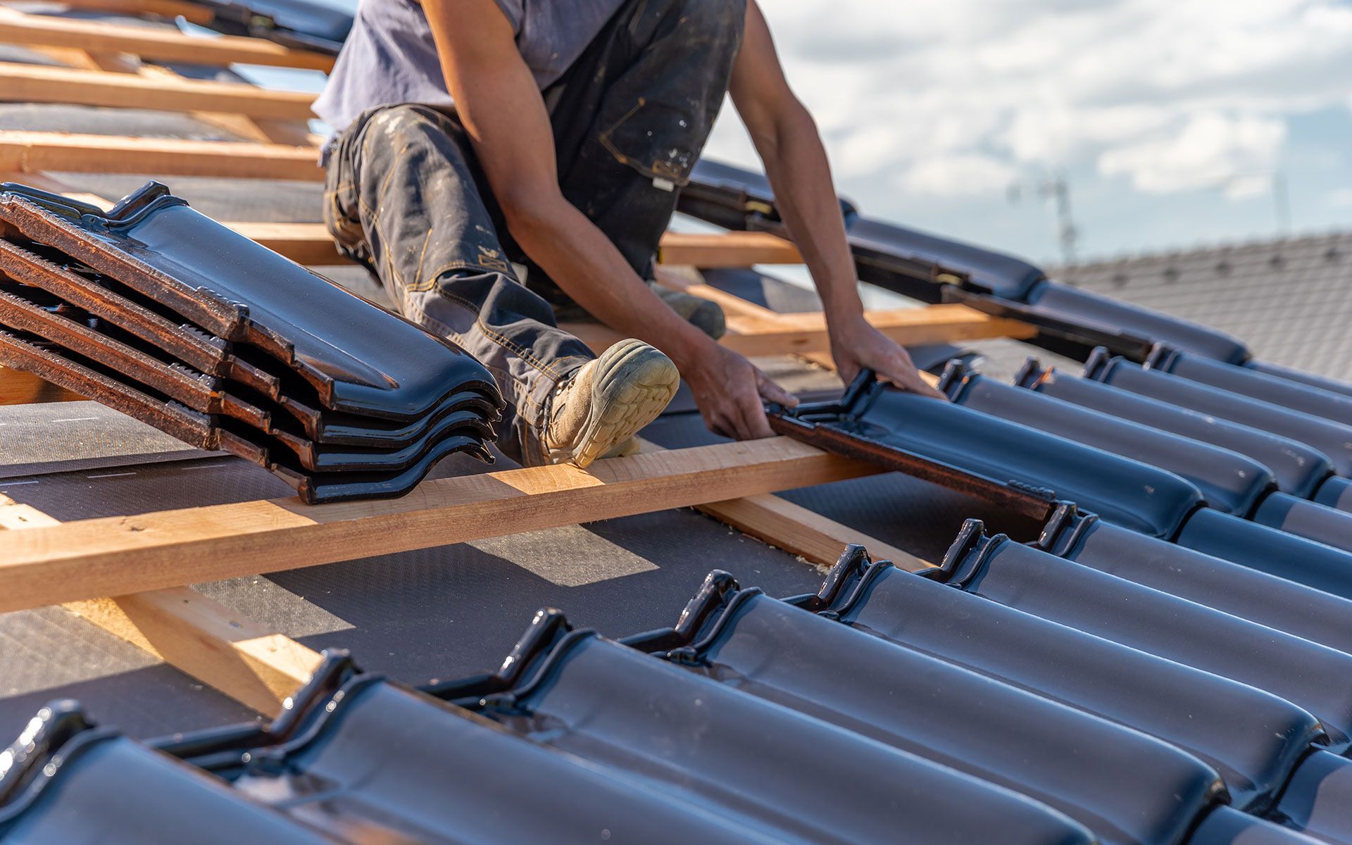 Roofer installing dark glazed tiles on a wooden roof structure against a cloudy sky.