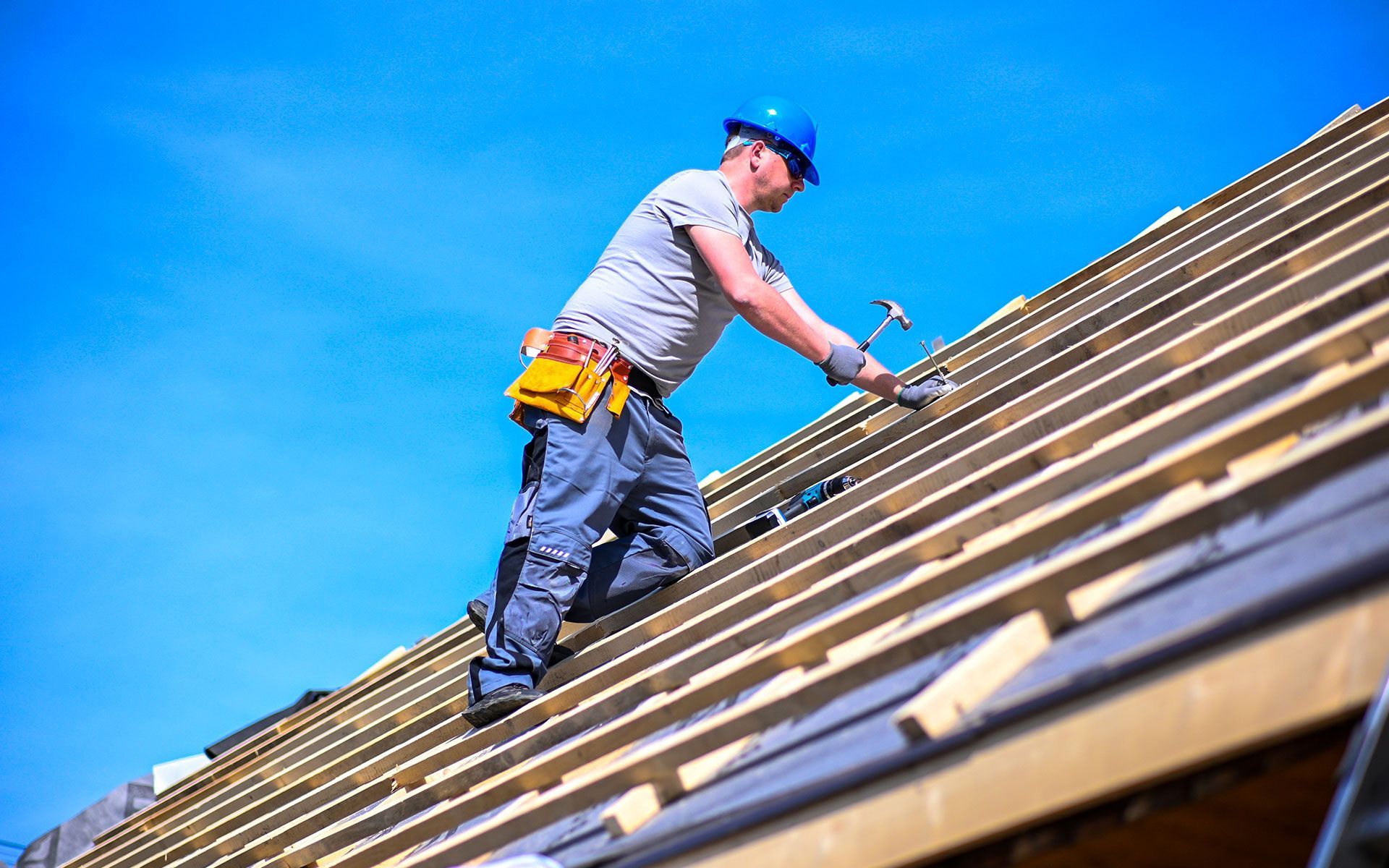 Roofer on a slanted roof wearing a blue hard hat, hammering wood under a clear blue sky.