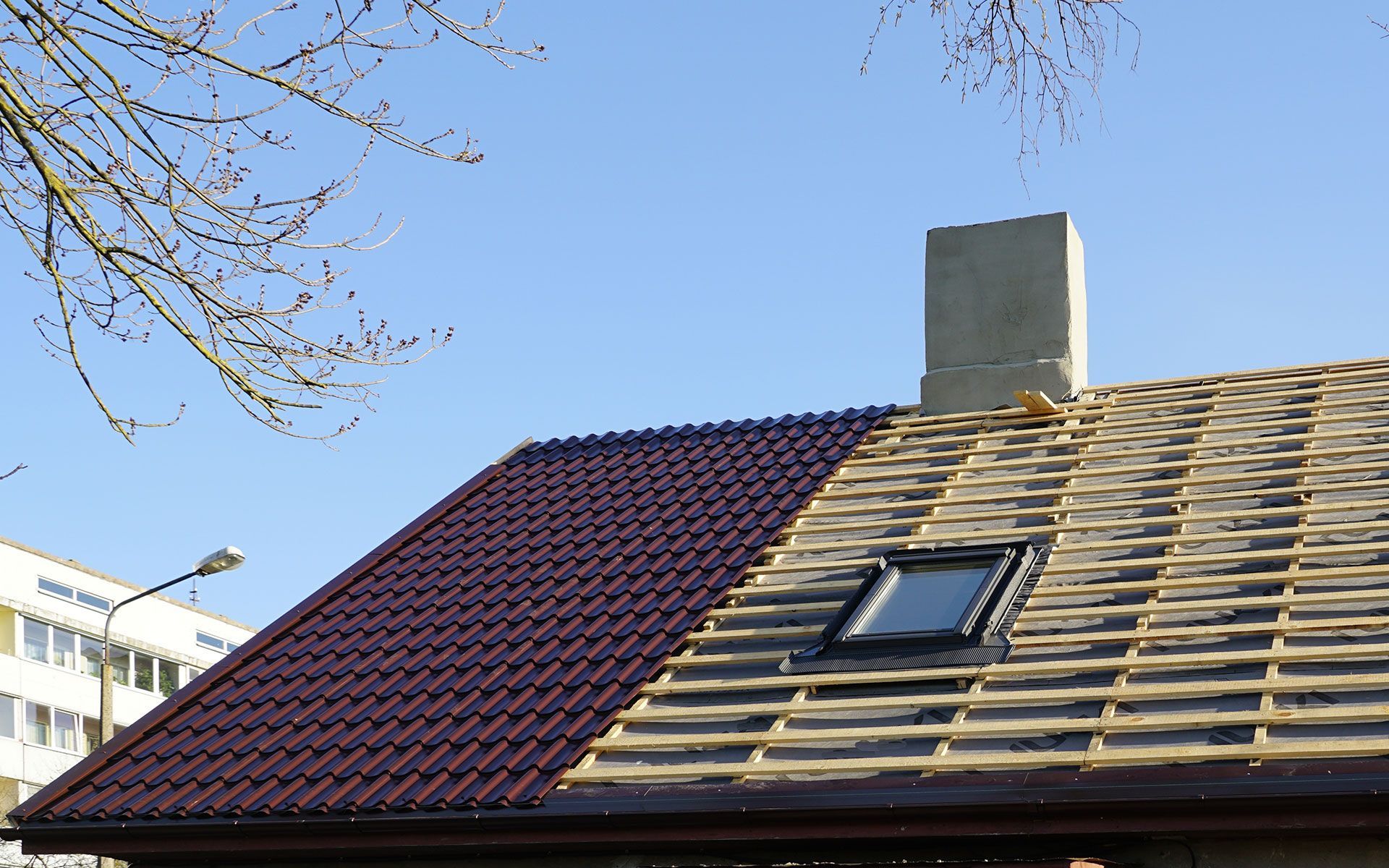Partially completed roof with dark red tiles, chimney, skylight, and blue sky.