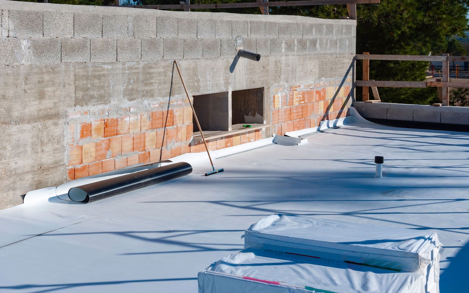 Rooftop construction with a white membrane, brick, and concrete walls. Black pipes, wood framing, and an open window.