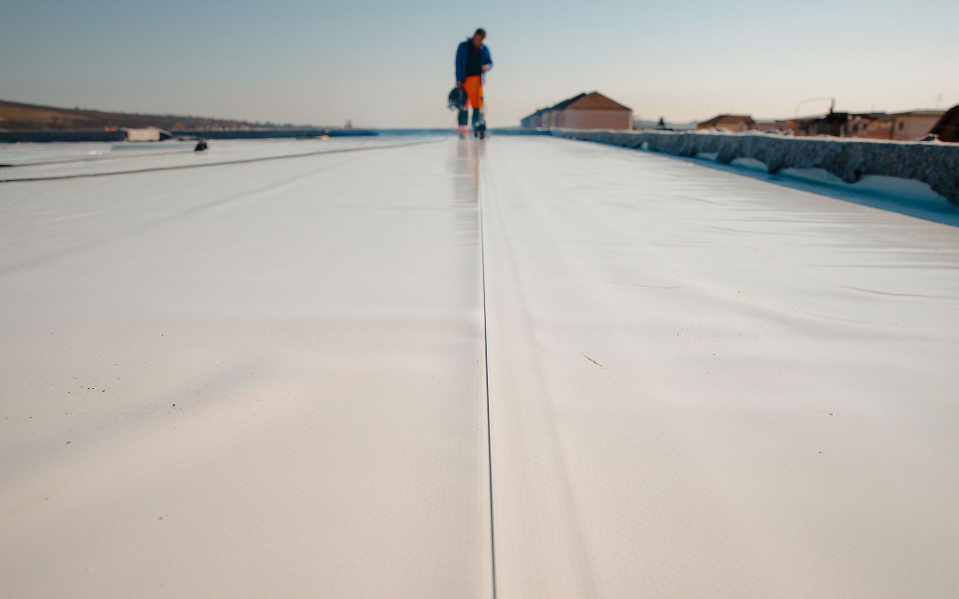 A worker on a white, flat roof, walking near a seam, with buildings in the background.