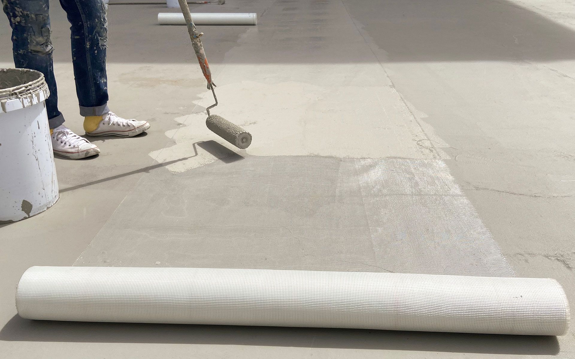 Person rolling sealant onto a concrete floor, next to a roll of mesh and a bucket.