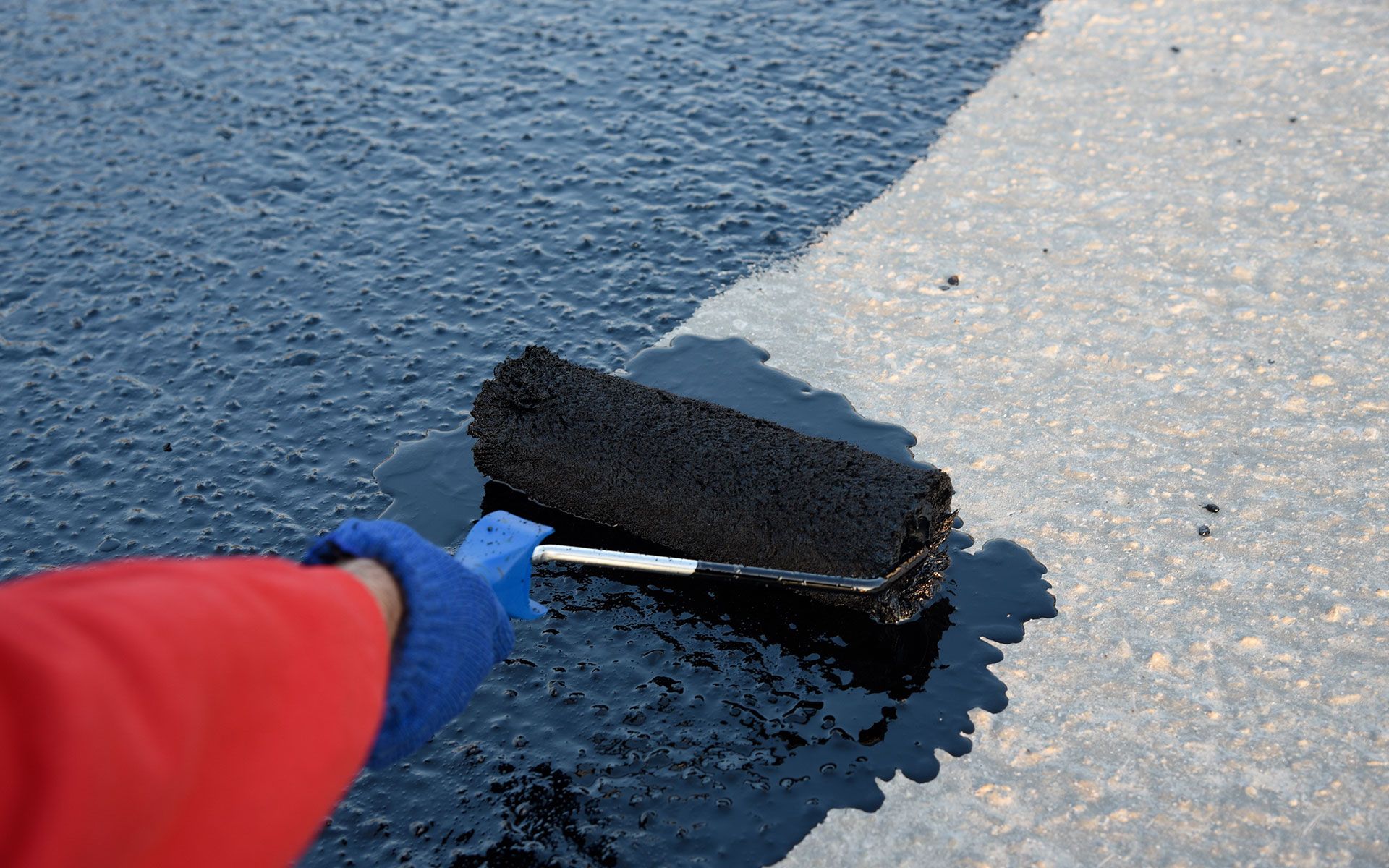 Person using a roller to apply black sealant to asphalt.