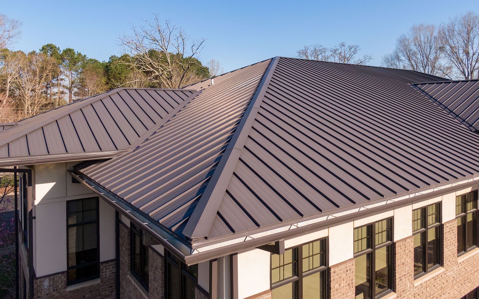Dark gray metal roof with angled ridges on a light brick building with windows.