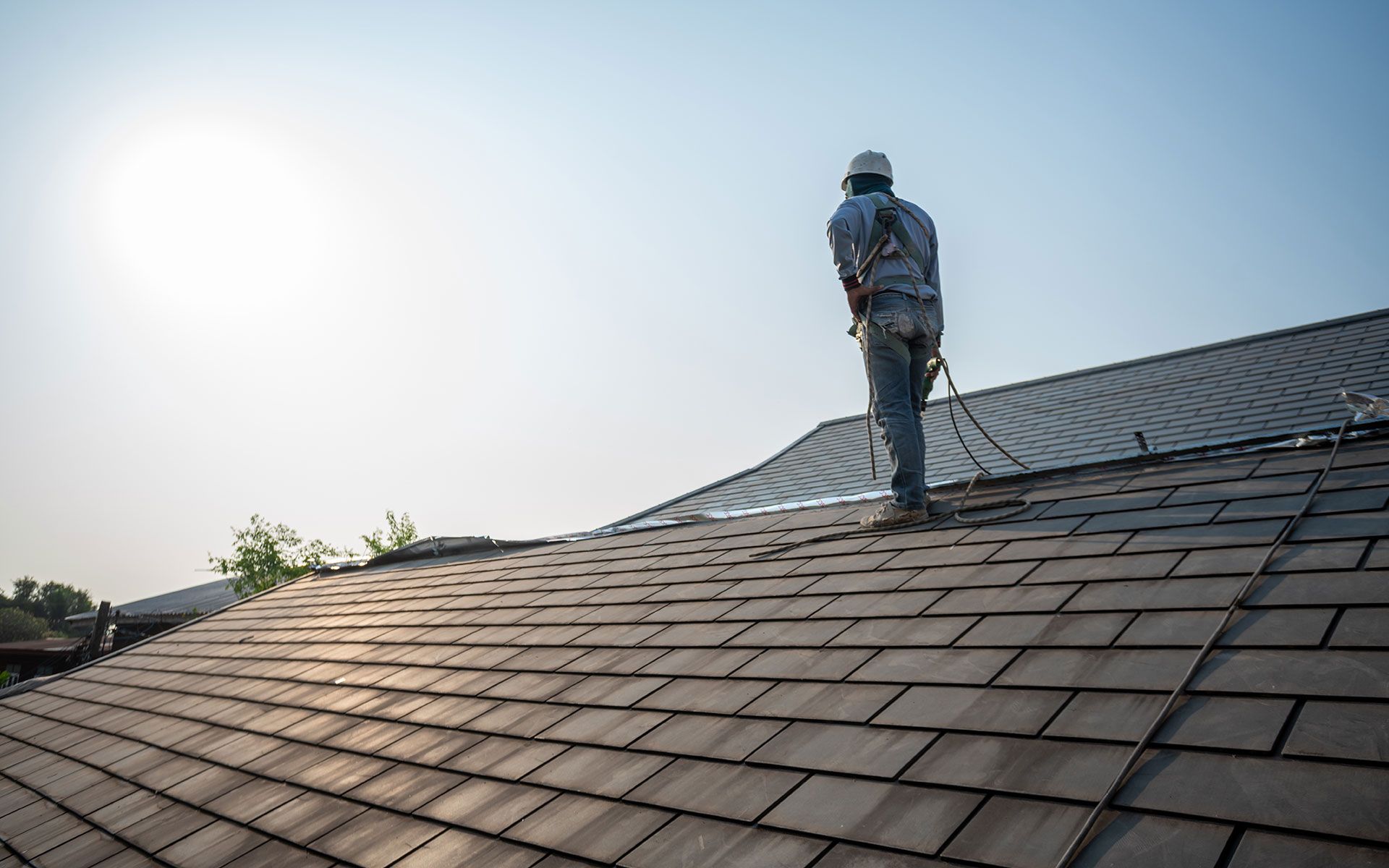 Roofer wearing a safety harness on a wood shingle roof under a bright sky.