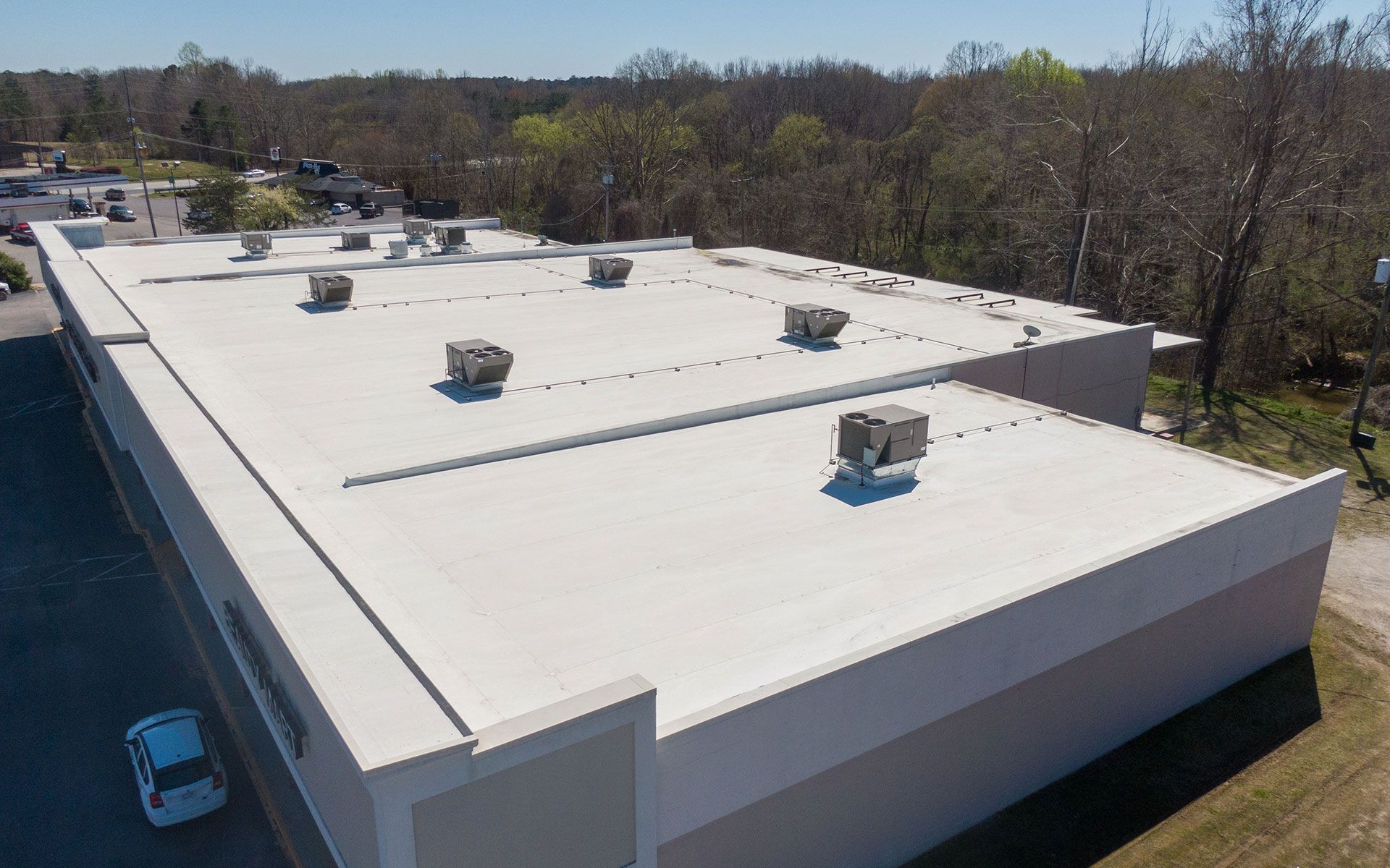 Aerial view of a large, white commercial building roof with HVAC units and surrounding trees.
