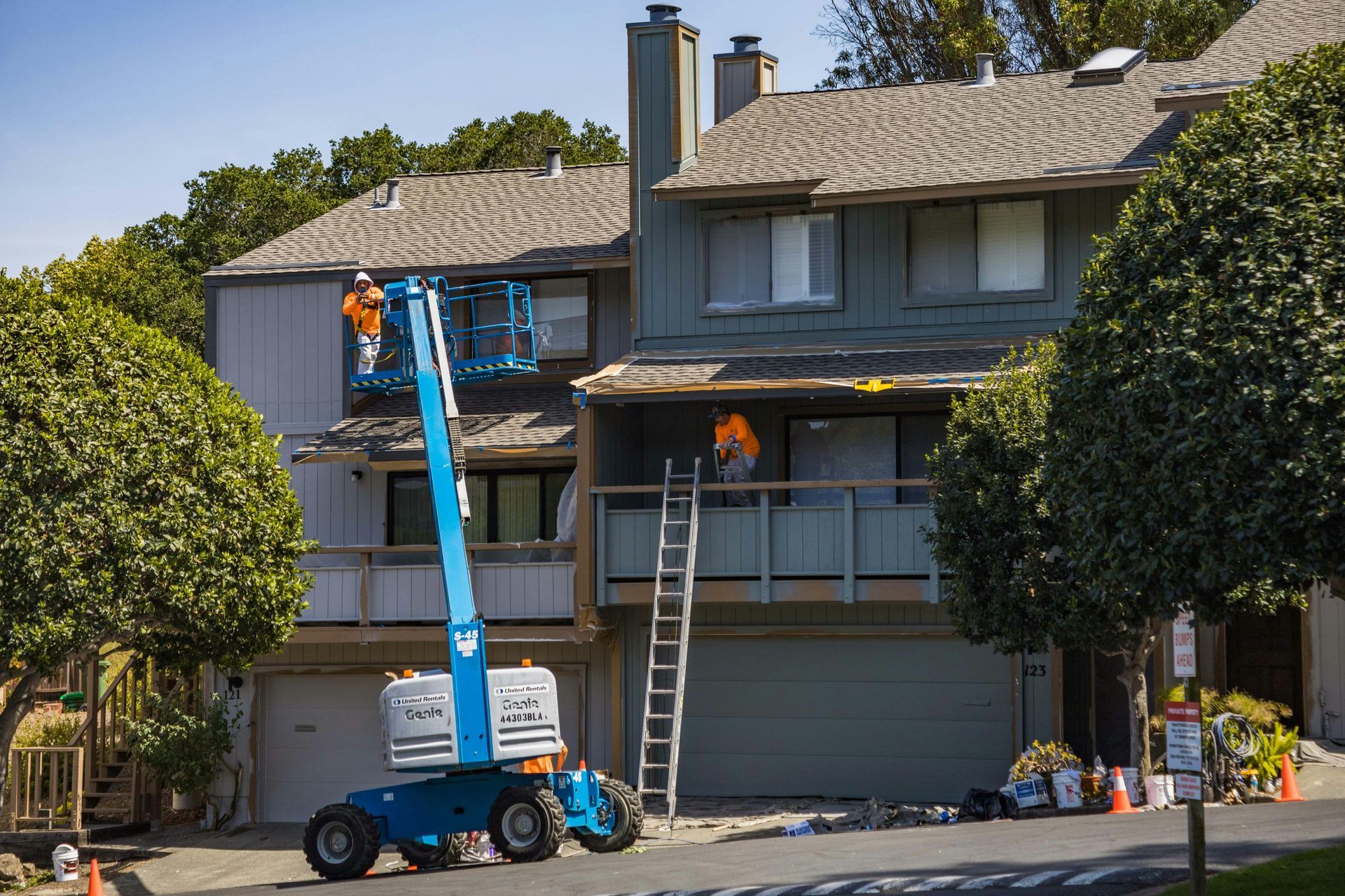 Two workers on lifts repairing the exterior of a two-story house with a ladder and garage below
