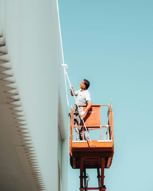 Worker on an orange lift painting a tall white building against a blue sky.