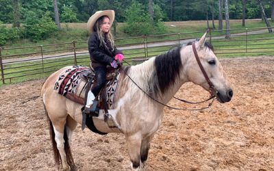 A child in a cowboy hat sits on a tan and white horse in a dirt riding arena with a fenced background.
