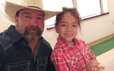A man in a cowboy hat and a child in a pink plaid shirt smile together in a well-lit indoor setting.