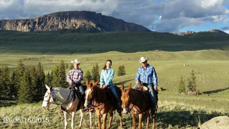 Three people wearing cowboy hats ride horses in a sunny, expansive landscape with mountains in the background.